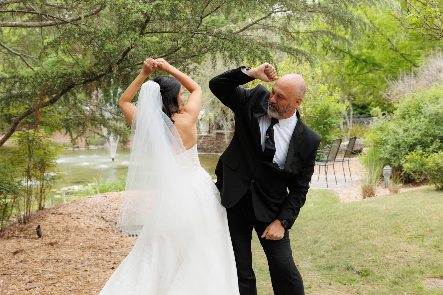 Bride and groom dancing outdoors at wedding, groom spinning bride in white gown and veil near trees.