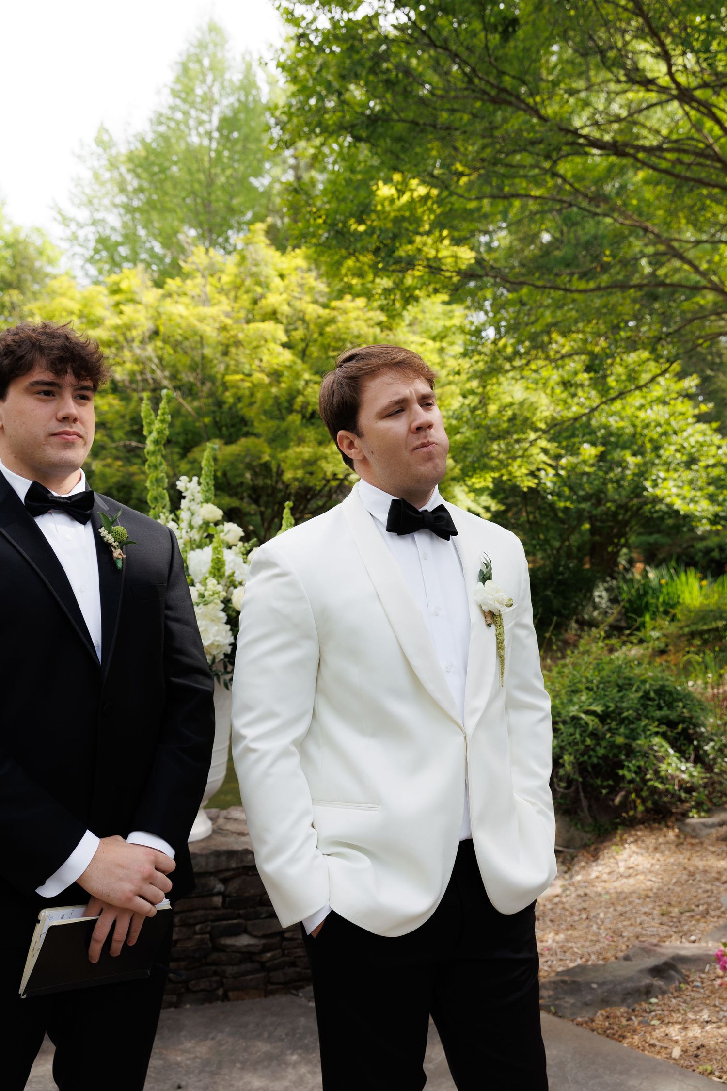 Groom in white tuxedo jacket with black bow tie stands outdoors with groomsman before wedding ceremony.