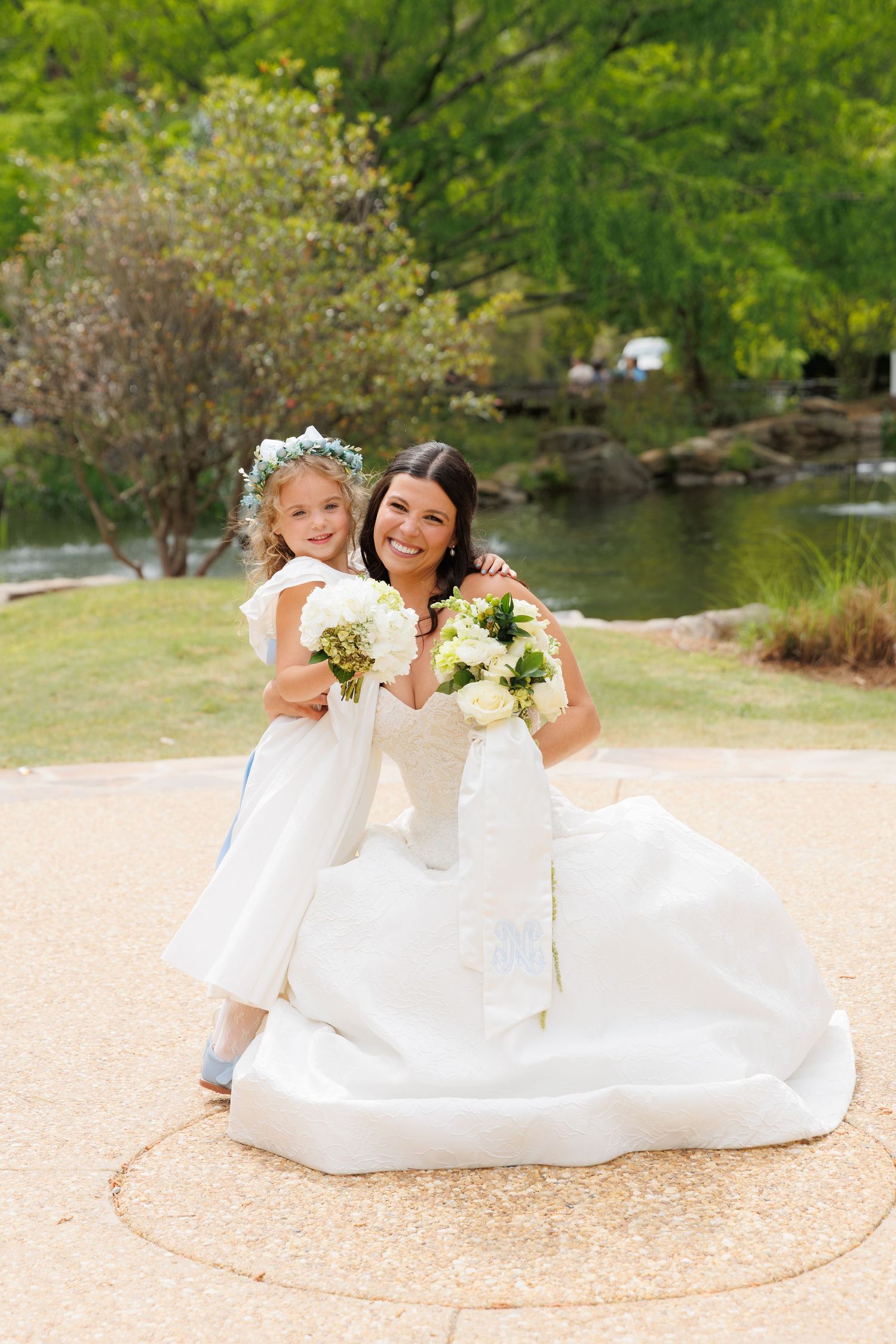 Smiling bride in white ball gown kneels beside flower girl wearing floral crown, both holding white bouquets outdoors.