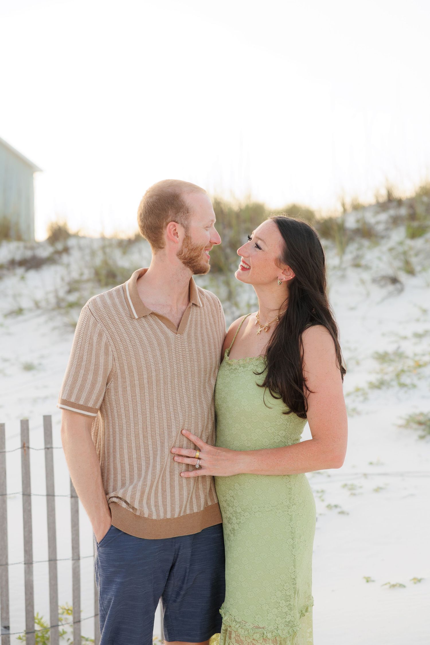 A happy couple embraces on a sandy beach at golden hour, woman in green dress, man in striped polo shirt.