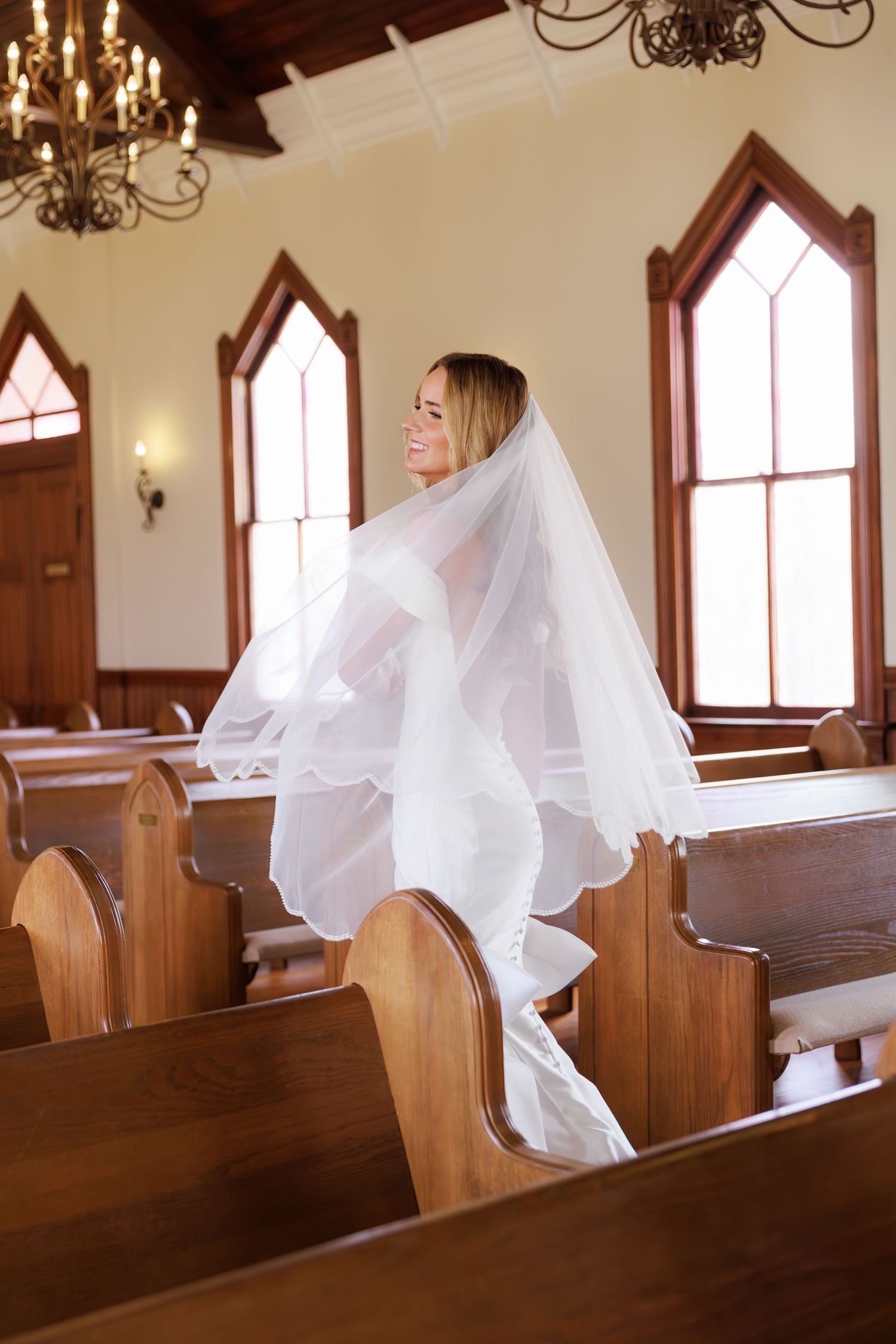 White tulle fabric drapes elegantly over wooden church pews in a sunlit chapel with arched Gothic windows and chandeliers.
