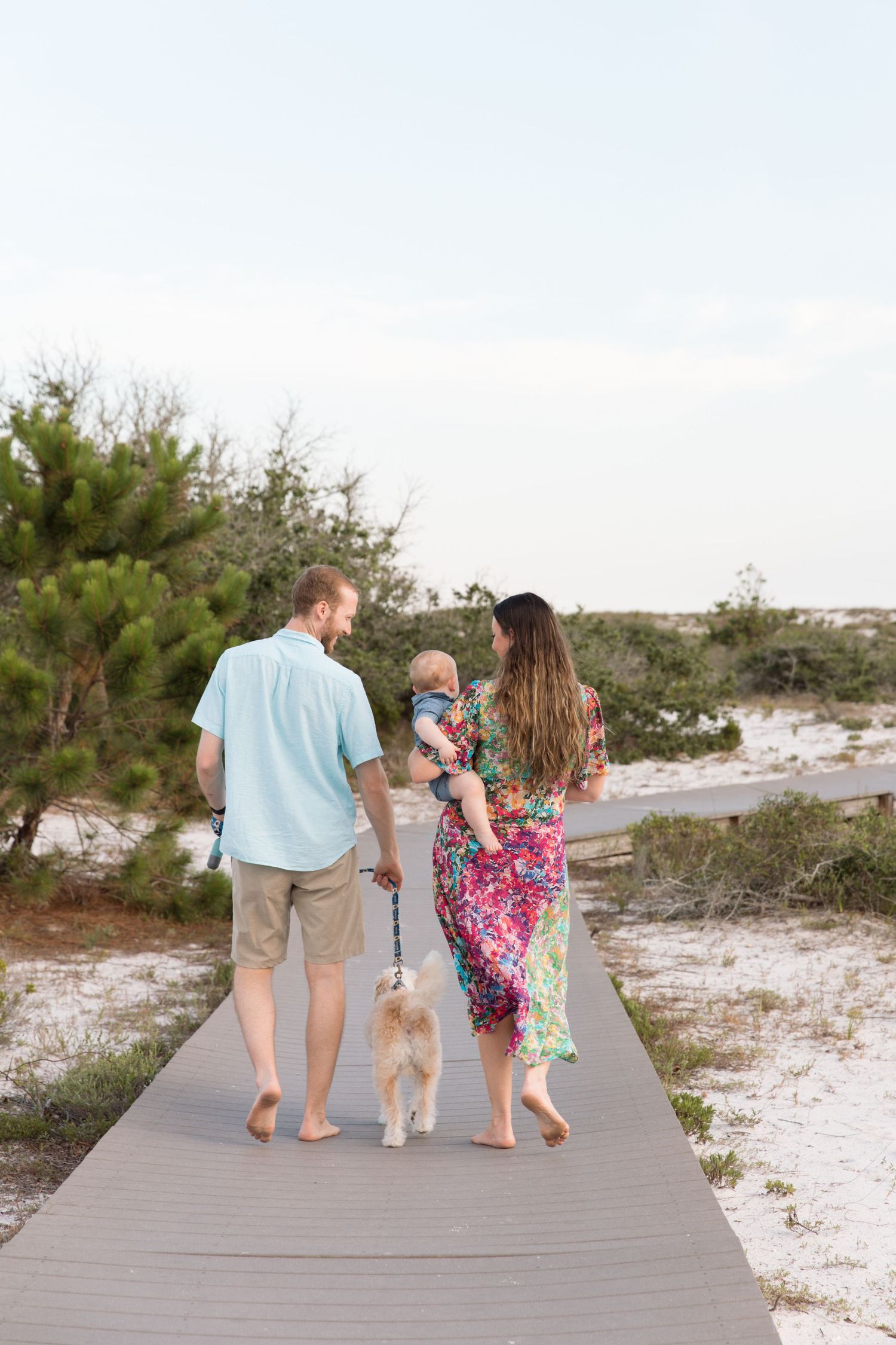 A family walks along a wooden boardwalk path through sandy dunes at the beach during sunset.