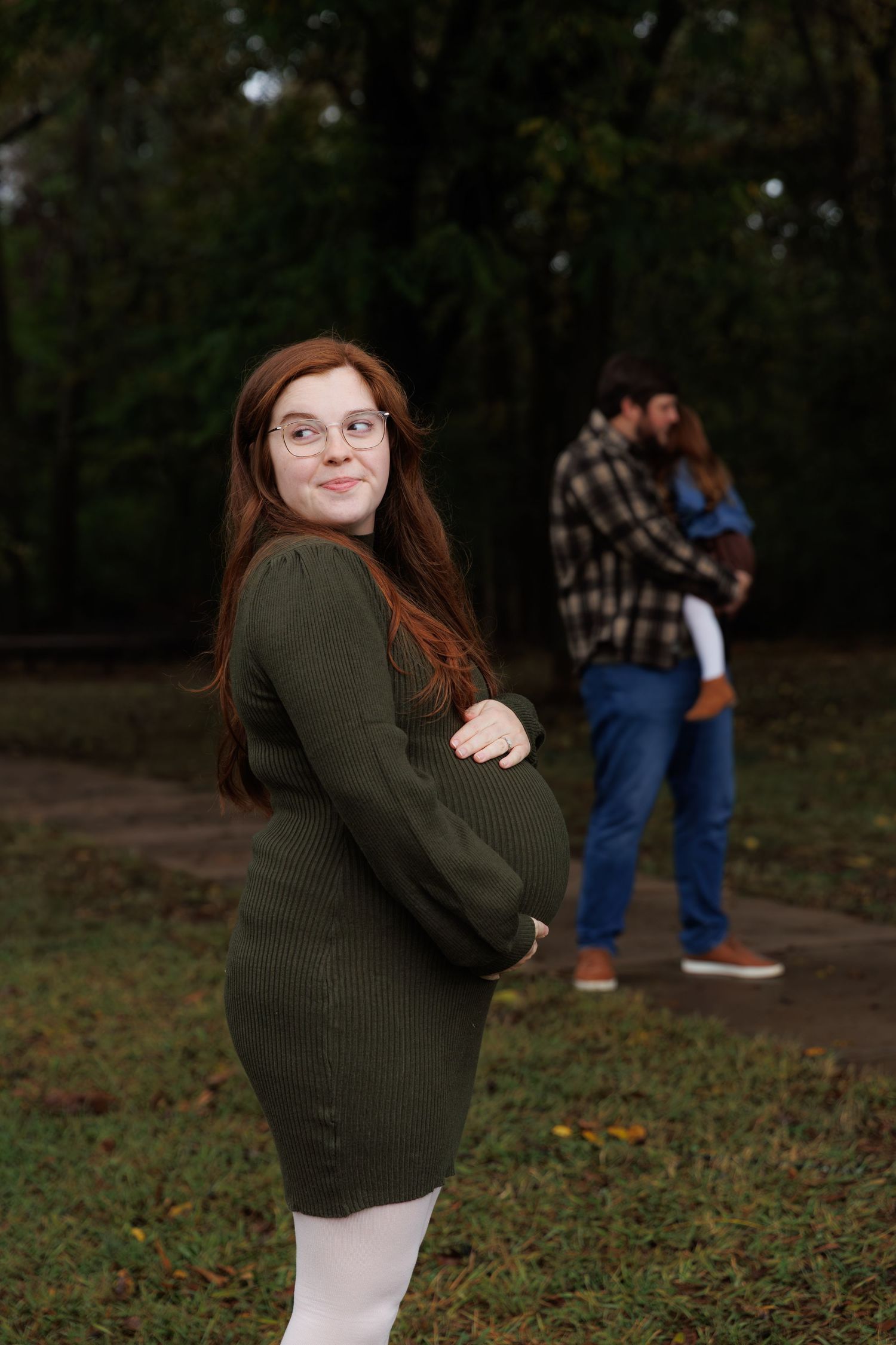 A person in a green sweater poses playfully in a park during autumn while others can be seen in the background.