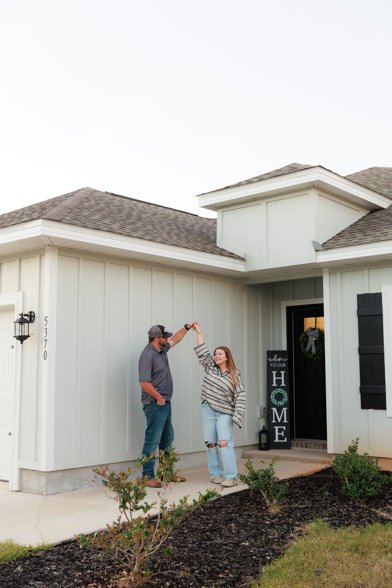 A couple celebrates joyfully on the front porch of their new white home with a decorative HOME sign by the door.