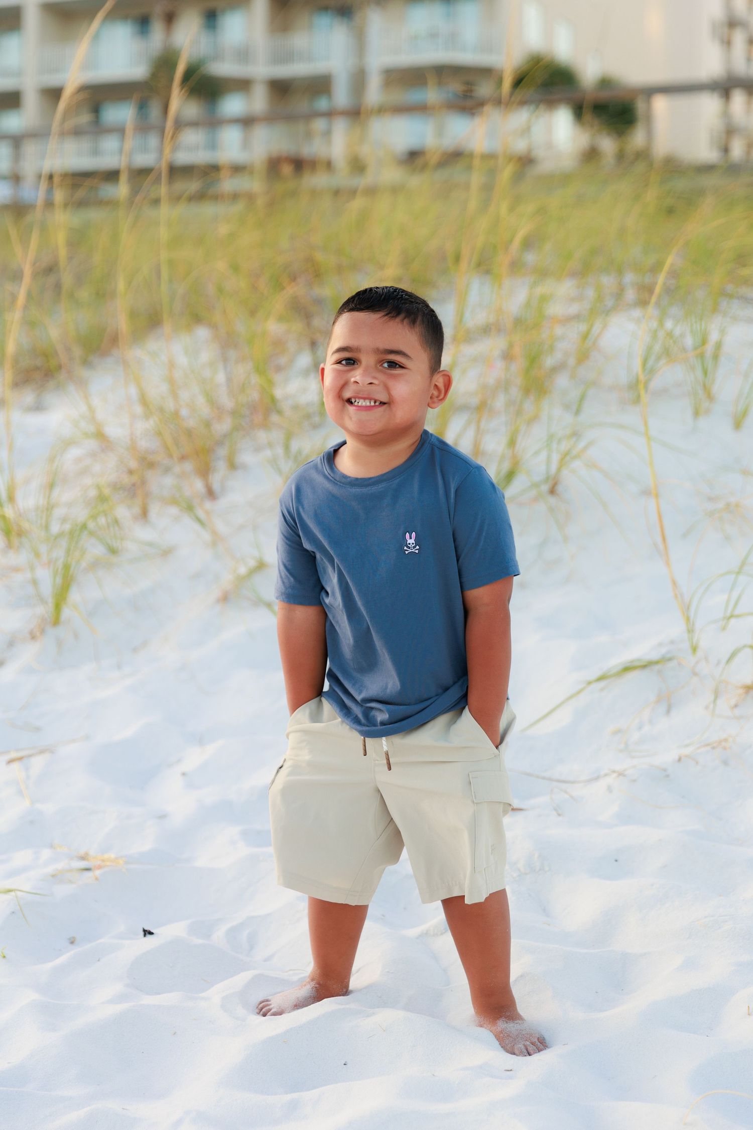 A young boy in a blue t-shirt and khaki shorts poses playfully on a white sandy beach.