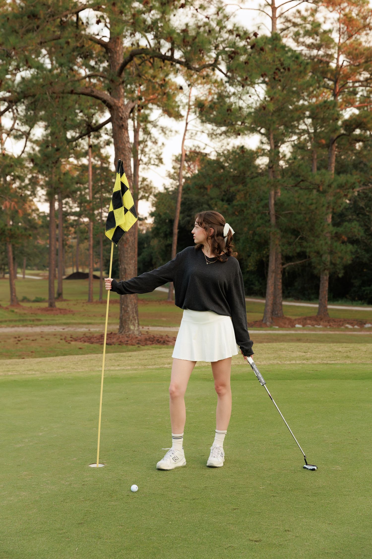 A golfer practicing putting on a green with pine trees in the background at sunset.