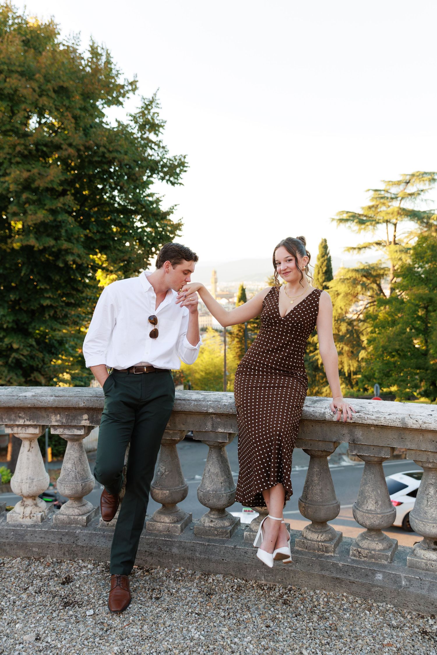 Elegant couple sharing a romantic moment on a stone balustrade terrace surrounded by lush green trees at golden hour.