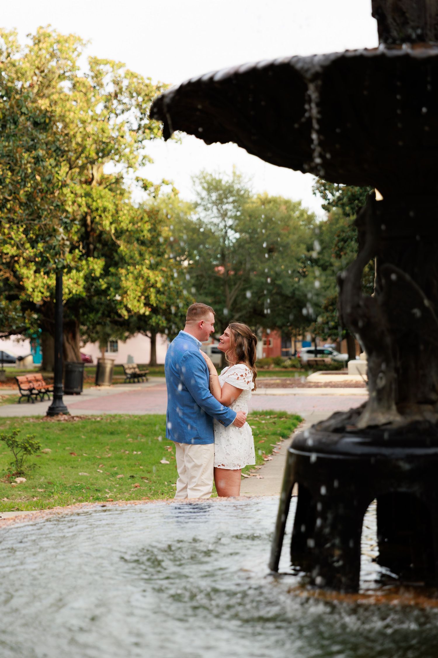 Couple embracing and smiling at each other beside a classic tiered fountain in an outdoor park setting.
