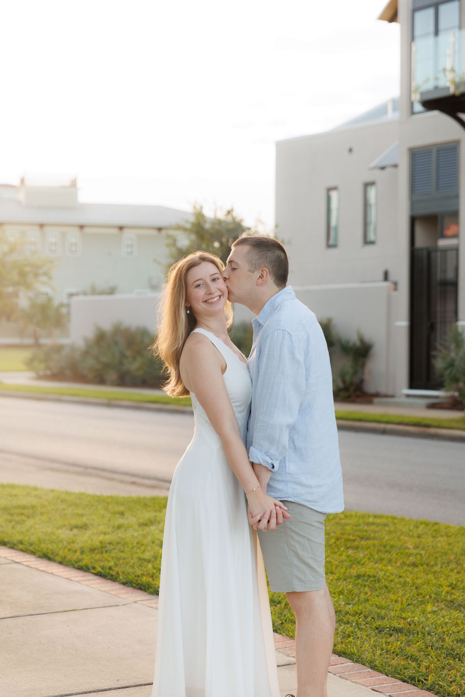 A couple shares a romantic moment outdoors at golden hour, woman in white dress smiling as man kisses her cheek.