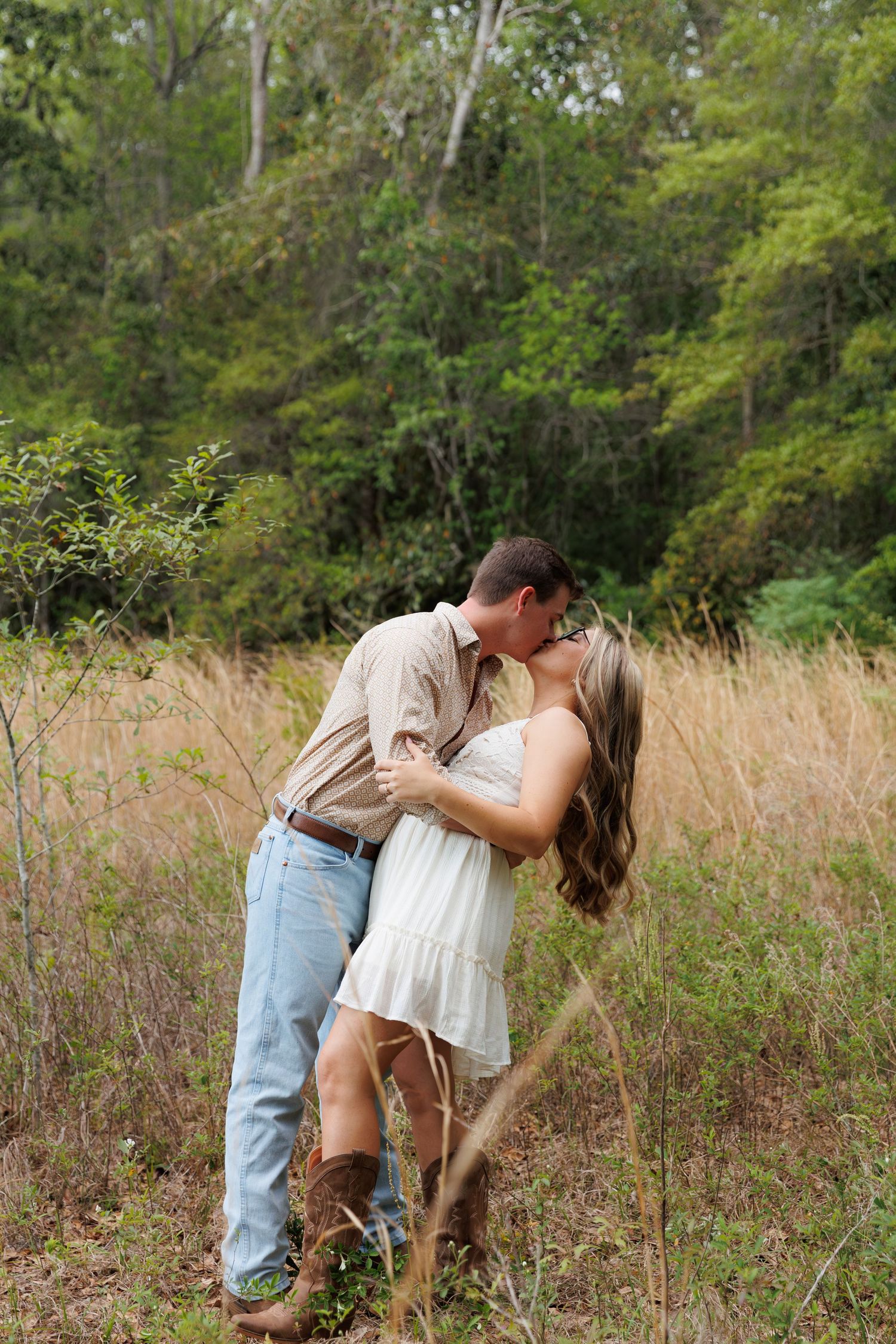 Couple sharing a romantic kiss in a golden field with lush green forest backdrop, woman in white dress and cowboy boots.