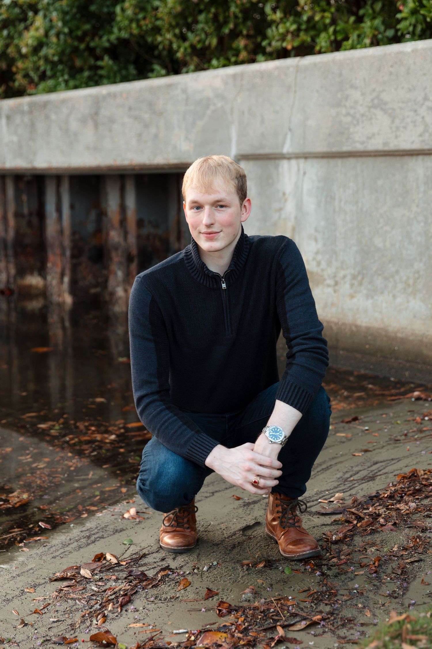 Young blond man in black sweater and jeans crouching outdoors on pavement surrounded by fallen leaves.