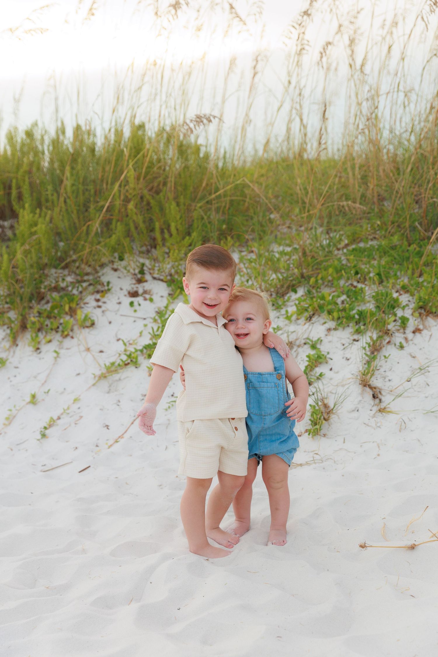 Two young children hugging on white sand beach dunes, boy in cream outfit and toddler in blue denim overalls.