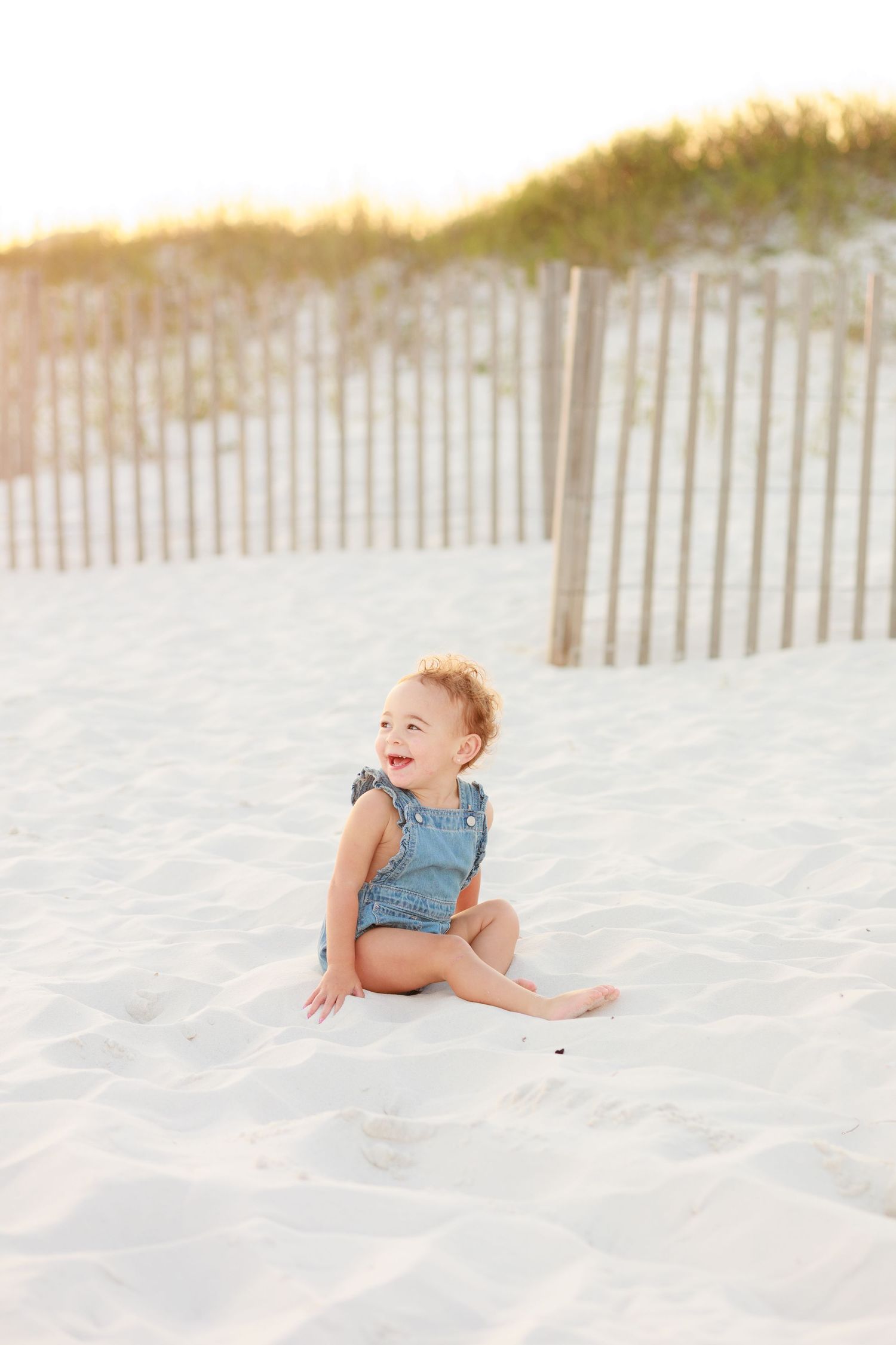 Happy baby in denim romper sitting on white sand beach laughing near wooden dune fence at sunset.