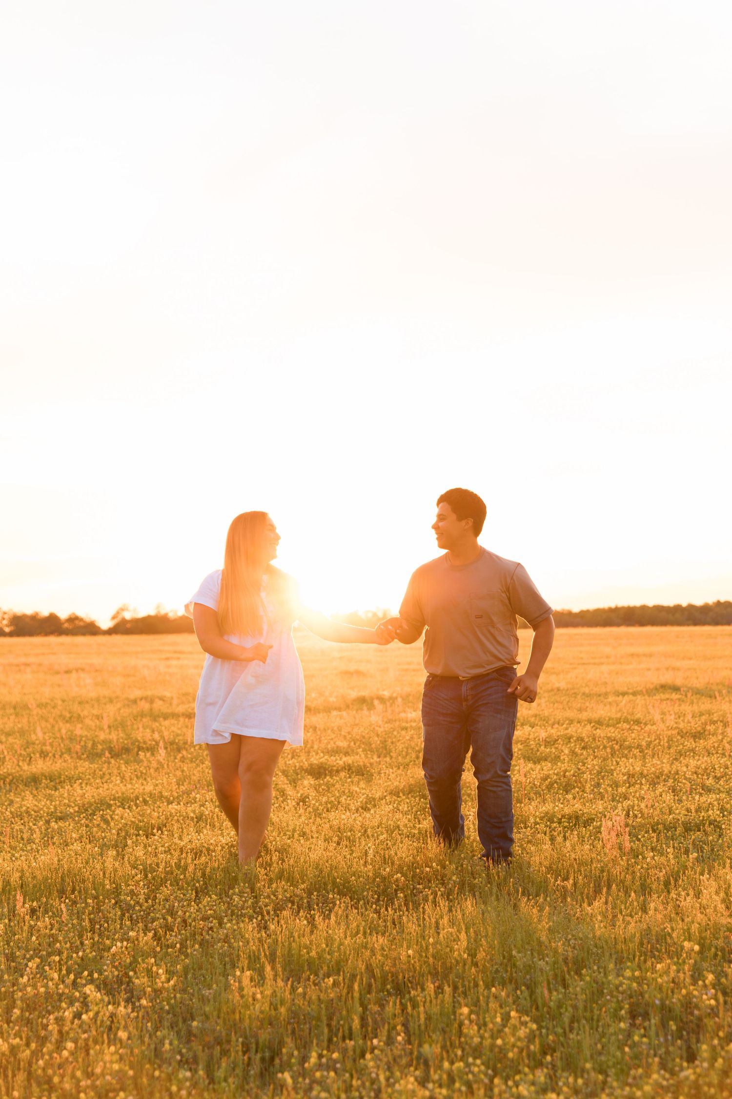 Couple standing in golden wheat field at sunset, woman in white dress and man in gray shirt enjoying romantic moment outdoors.