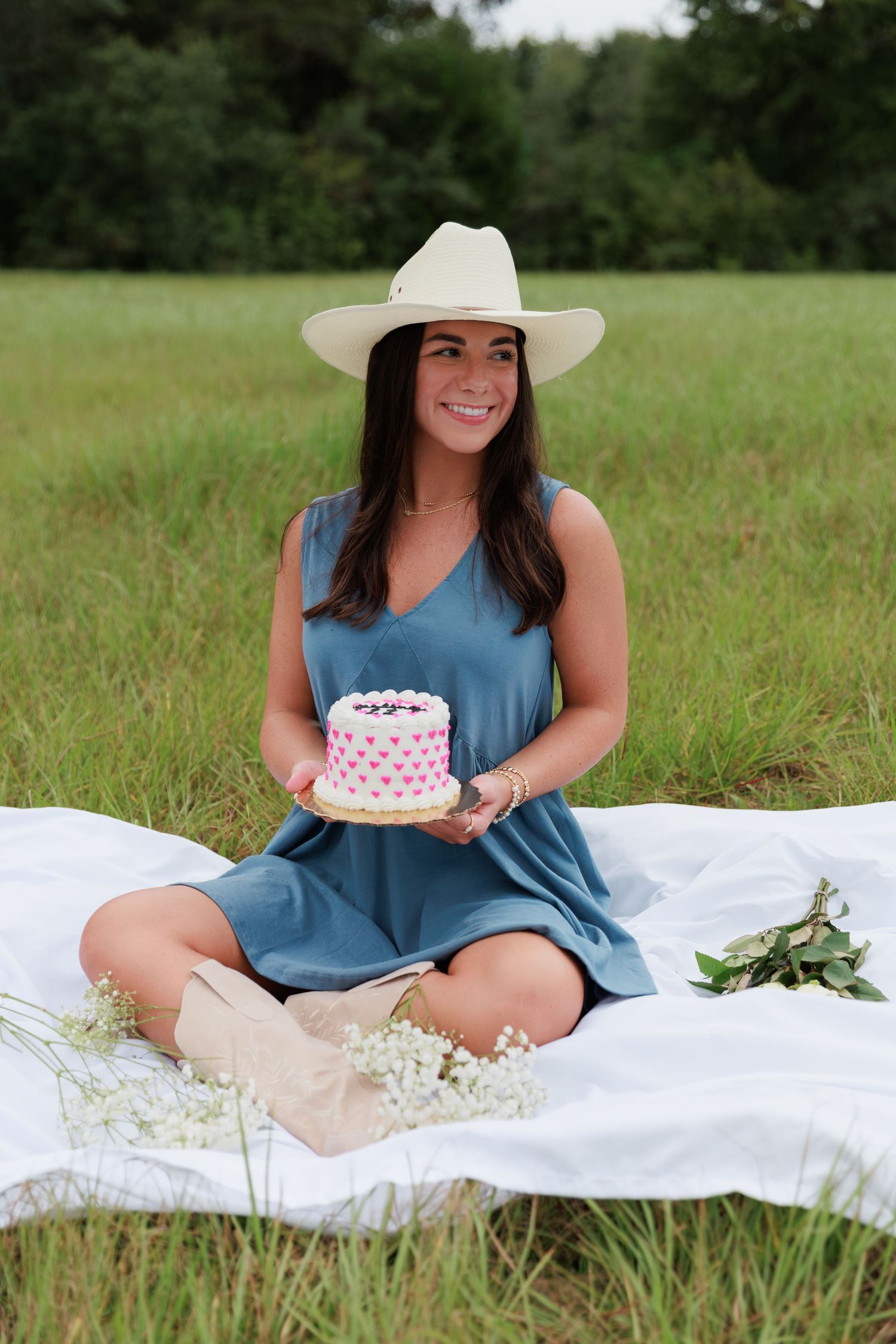 Outdoor celebration photo with small pink cake, cowboy hat and casual blue dress on white blanket.