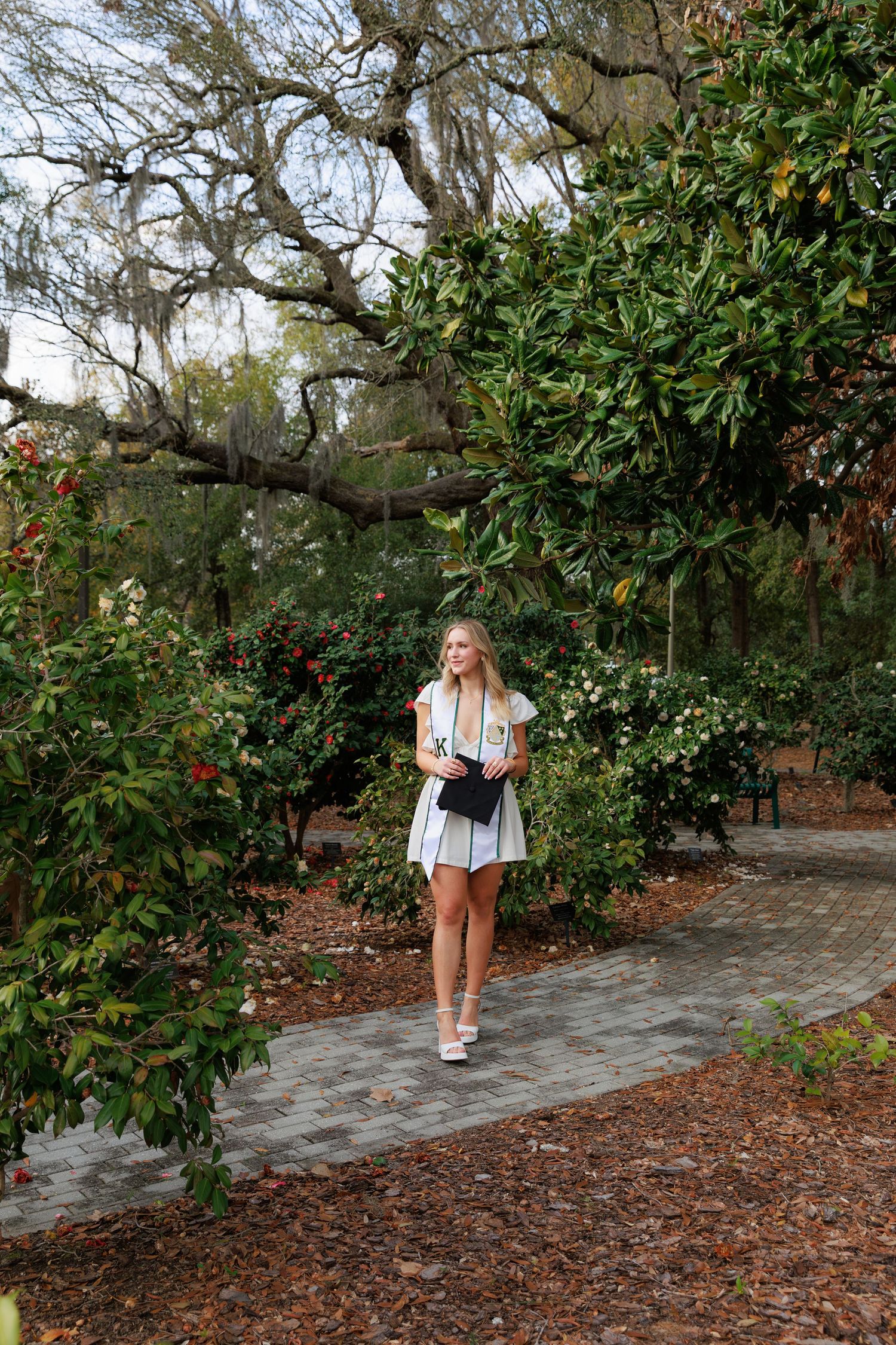 Blonde woman in striped dress walking on brick path through lush green garden with flowering shrubs.