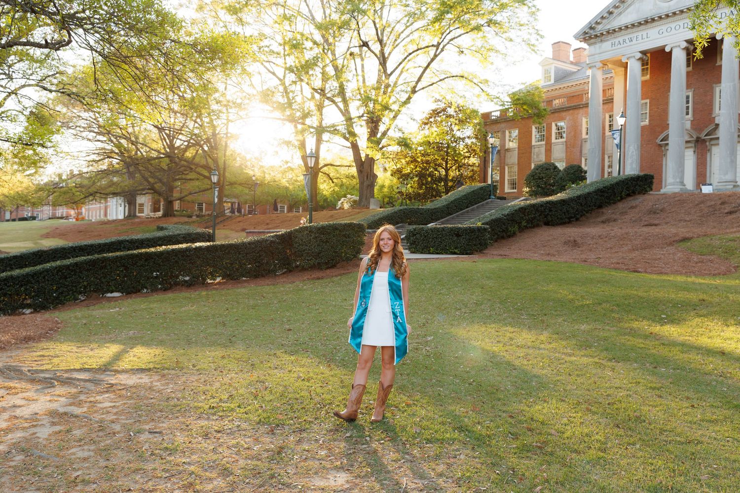 Young woman in teal dress and cowboy boots standing on green campus lawn at golden hour near historic brick building.