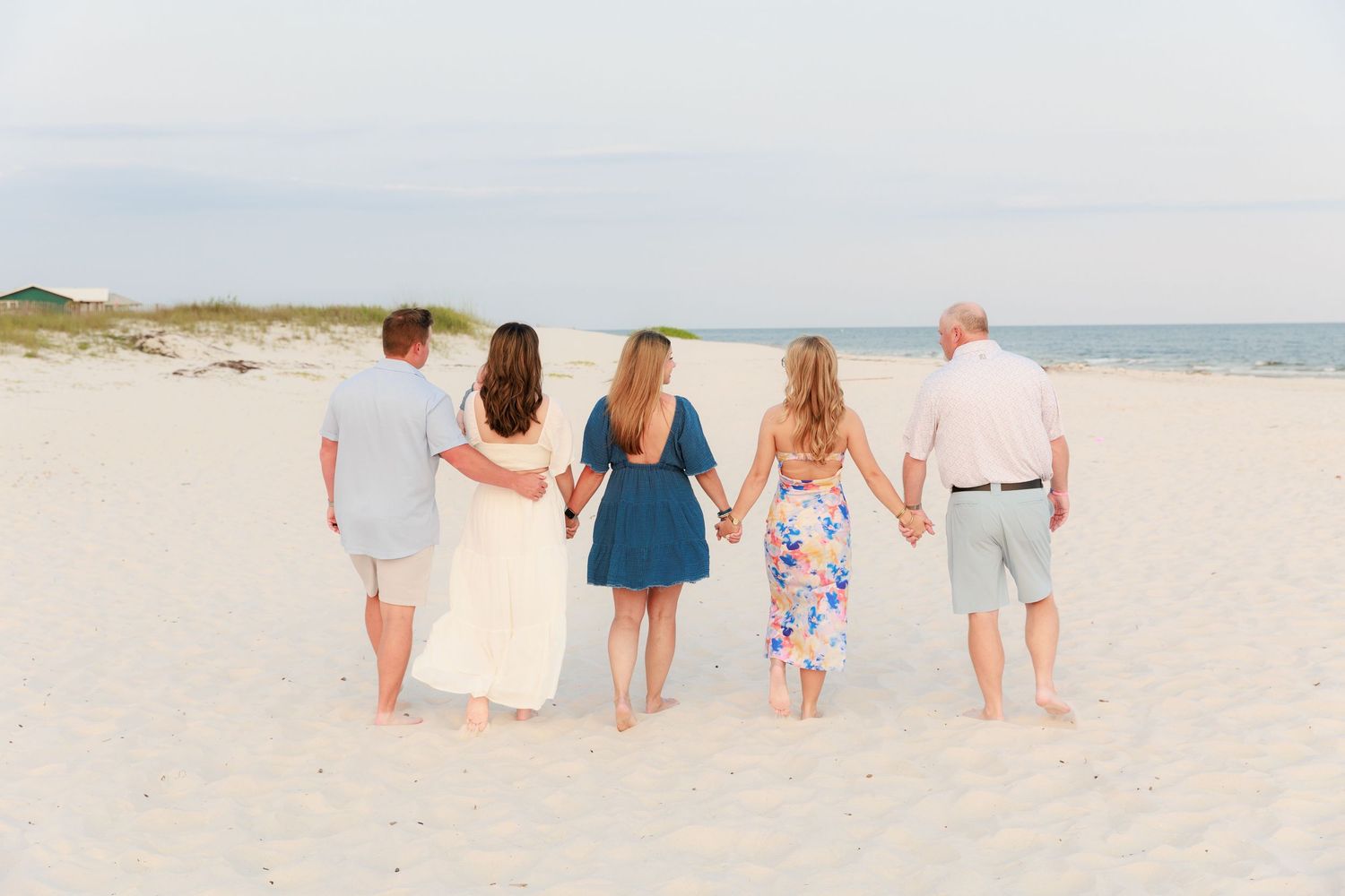 Family of five walking hand-in-hand along sandy beach toward ocean at sunset.