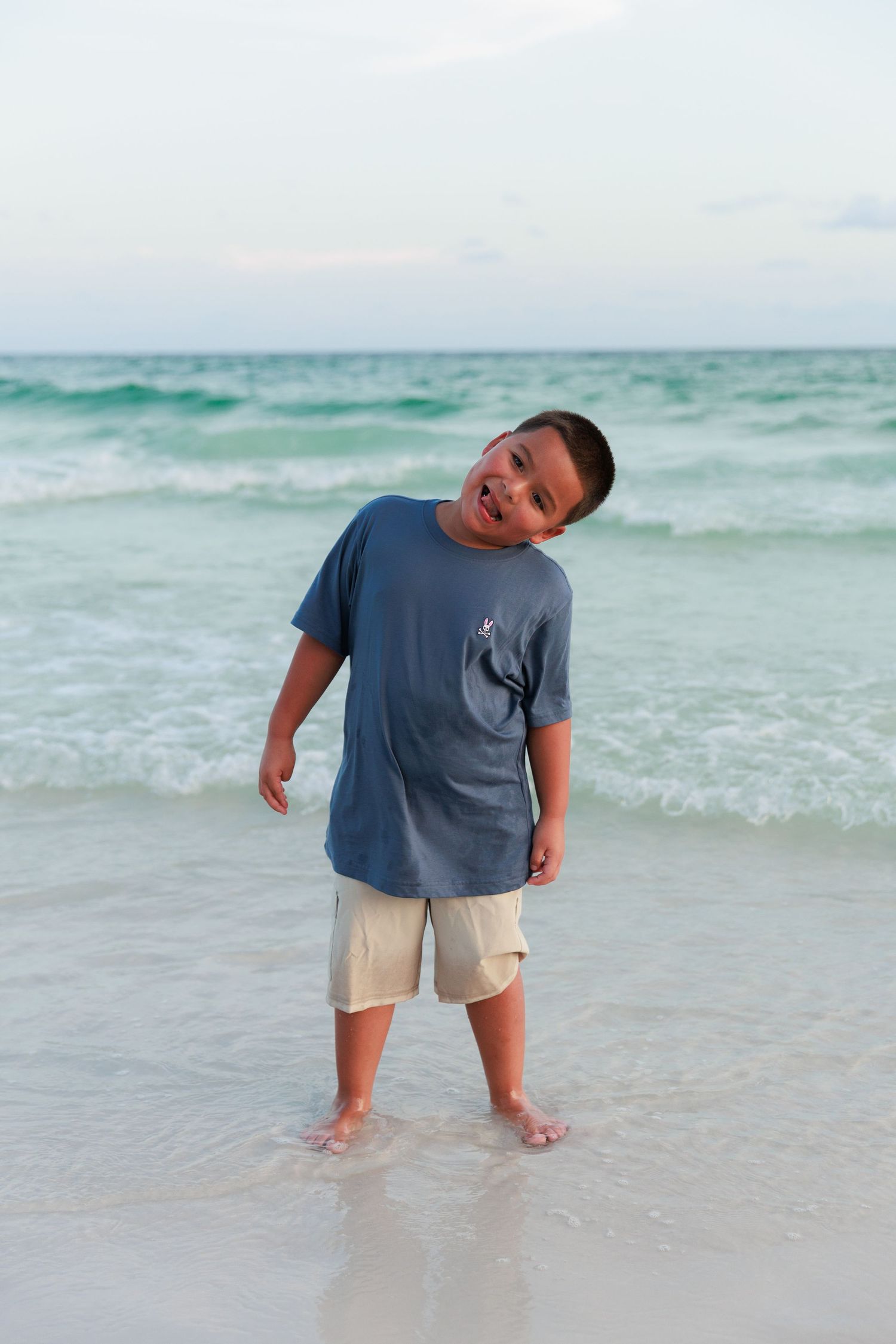 A person in a blue t-shirt and light shorts plays in the shallow ocean water at the beach.