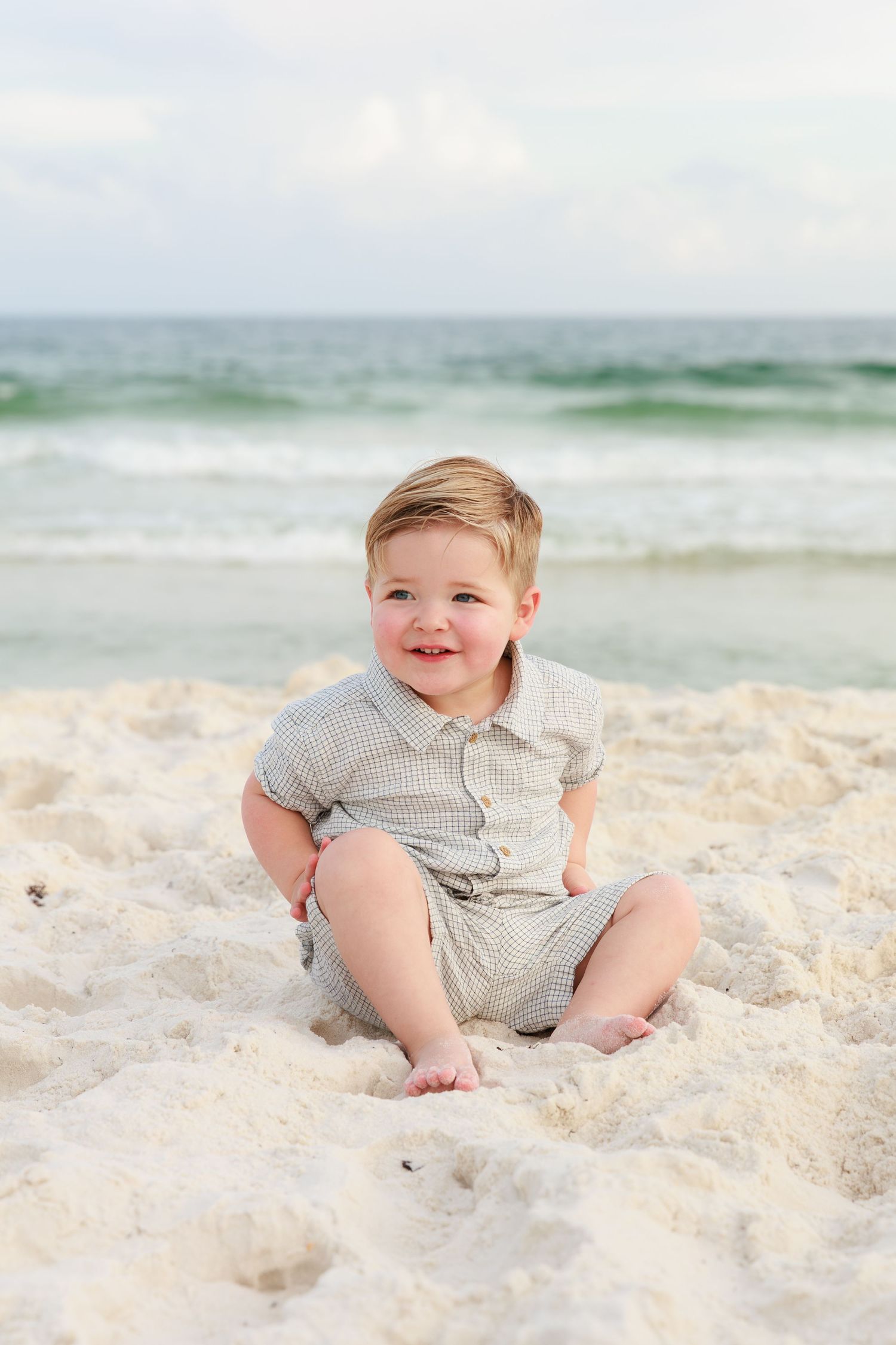 A baby in gray clothing sits and plays on pristine white sand at the beach with ocean waves in the background.