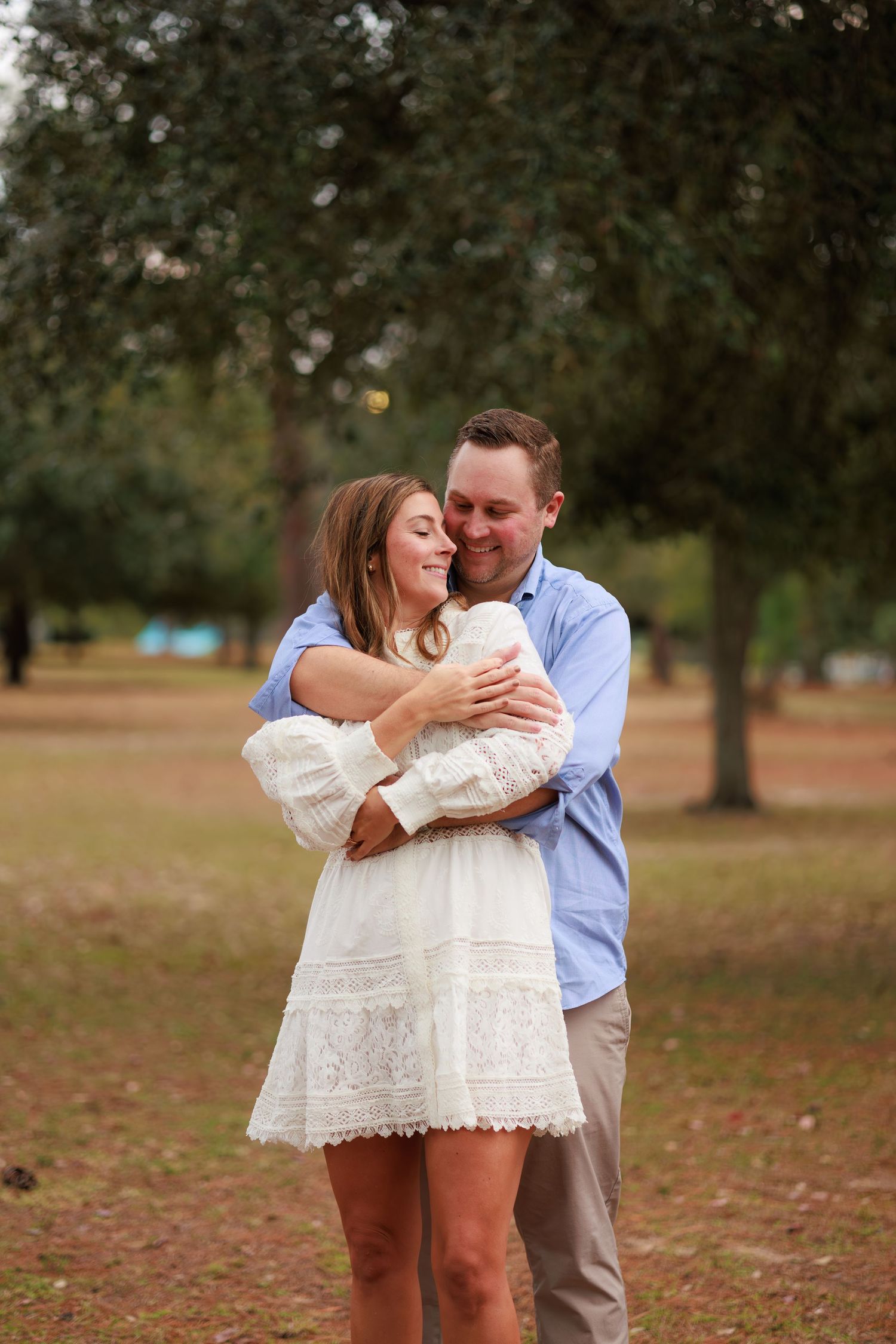 A couple embraces outdoors in a park with autumn leaves on the ground and trees in the background.
