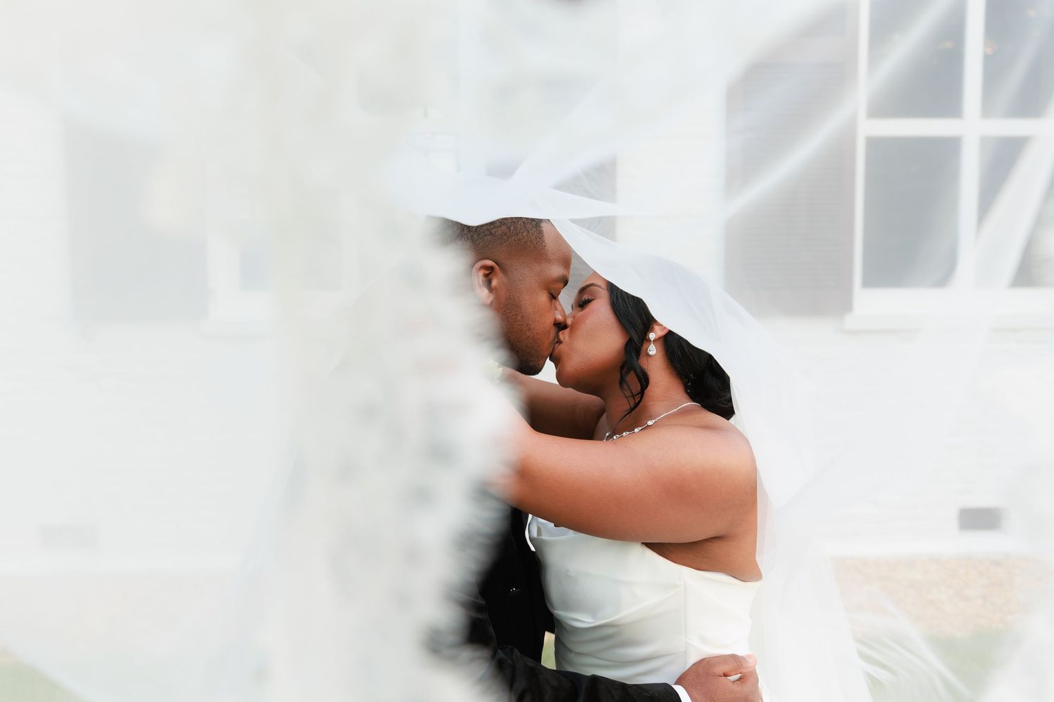 A romantic wedding couple embraces and shares a tender kiss in a light-filled venue with white decor.