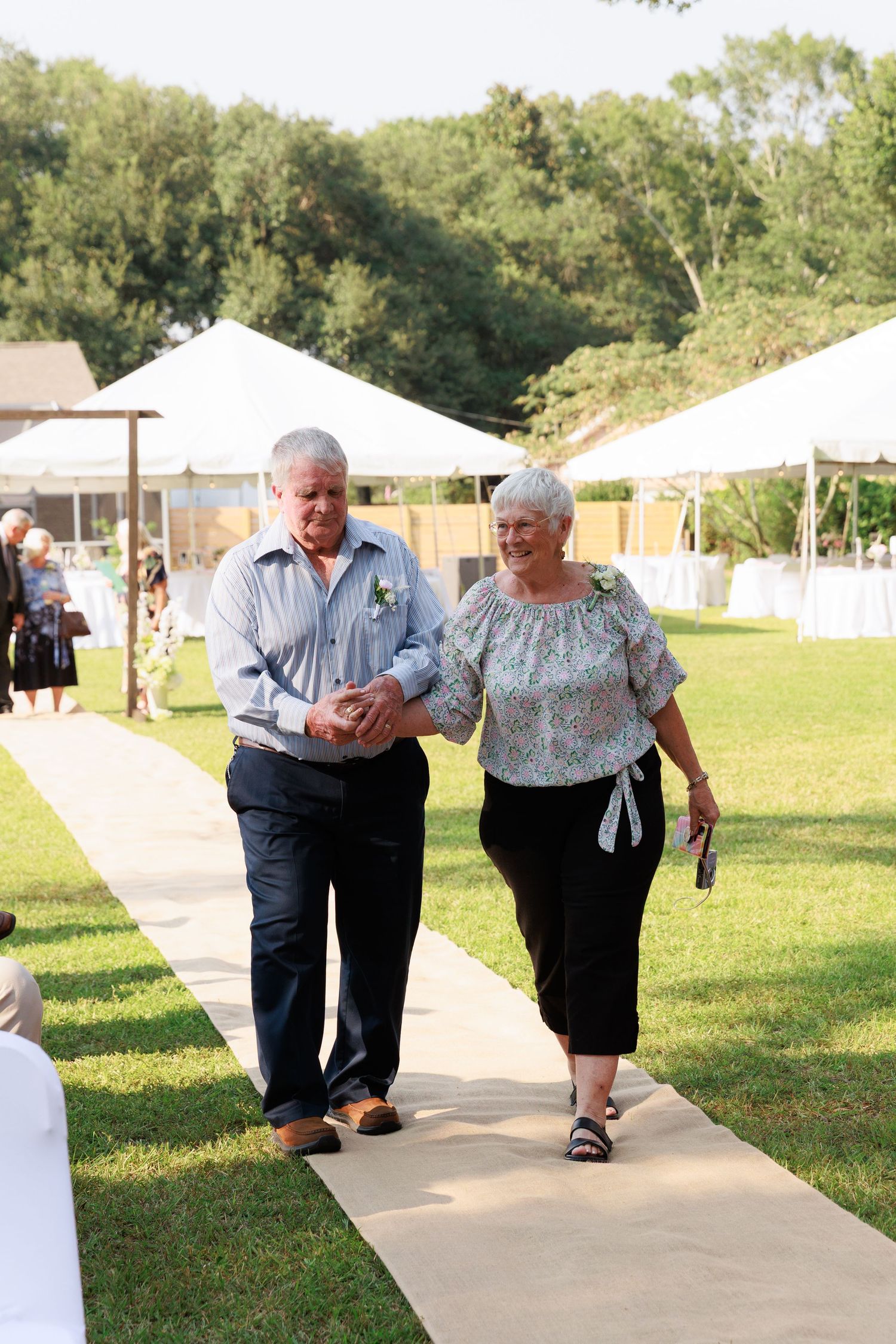 A paved pathway winds through a sunny grassy area with white canopy tents and trees visible.