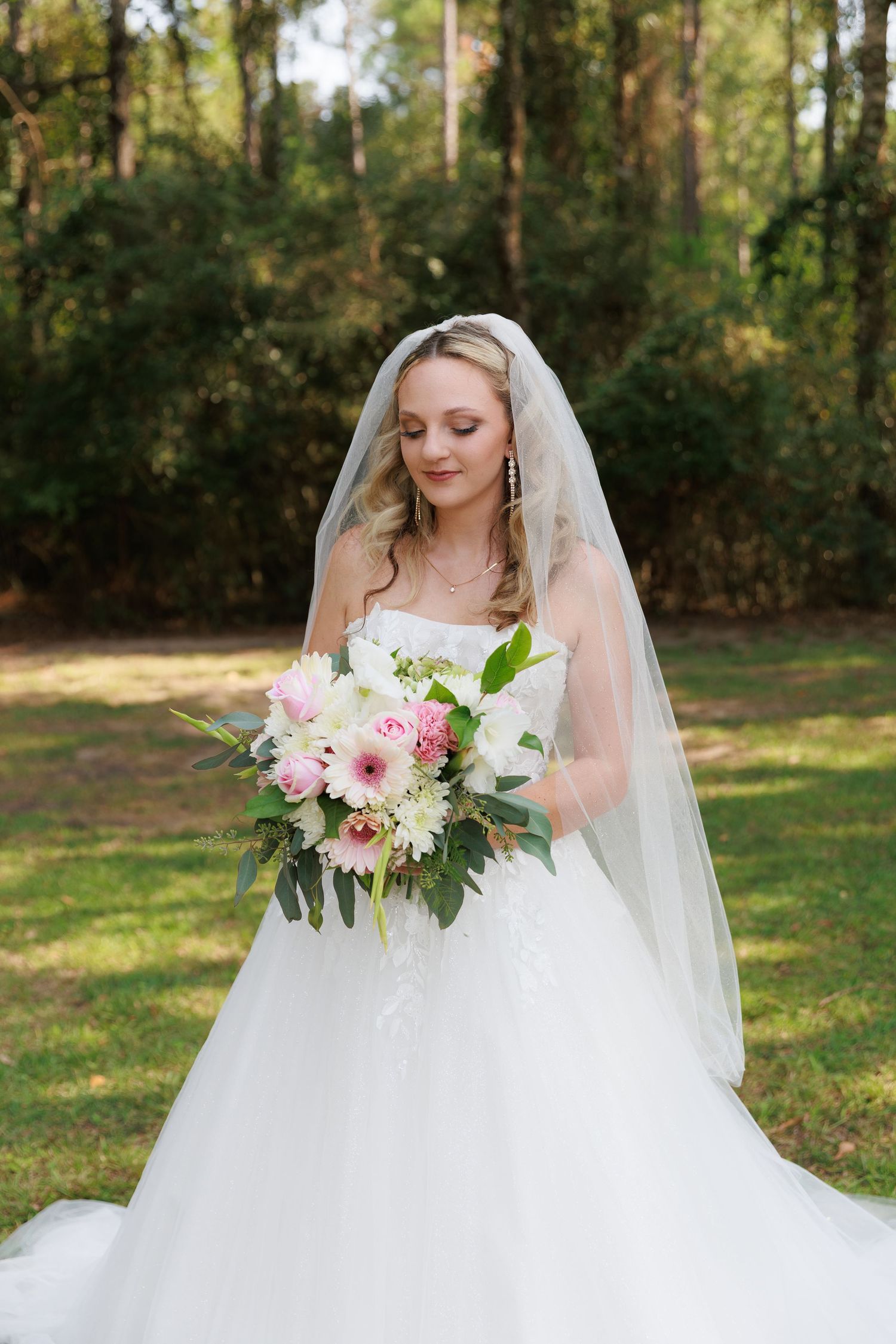 A bride in a flowing white wedding gown holds a pink and white floral bouquet in an outdoor garden setting.