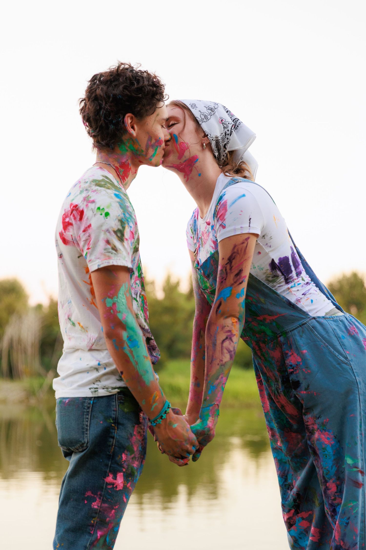 Couple splattered with vibrant paint powder hold hands and lean towards each other near a lakeside setting.