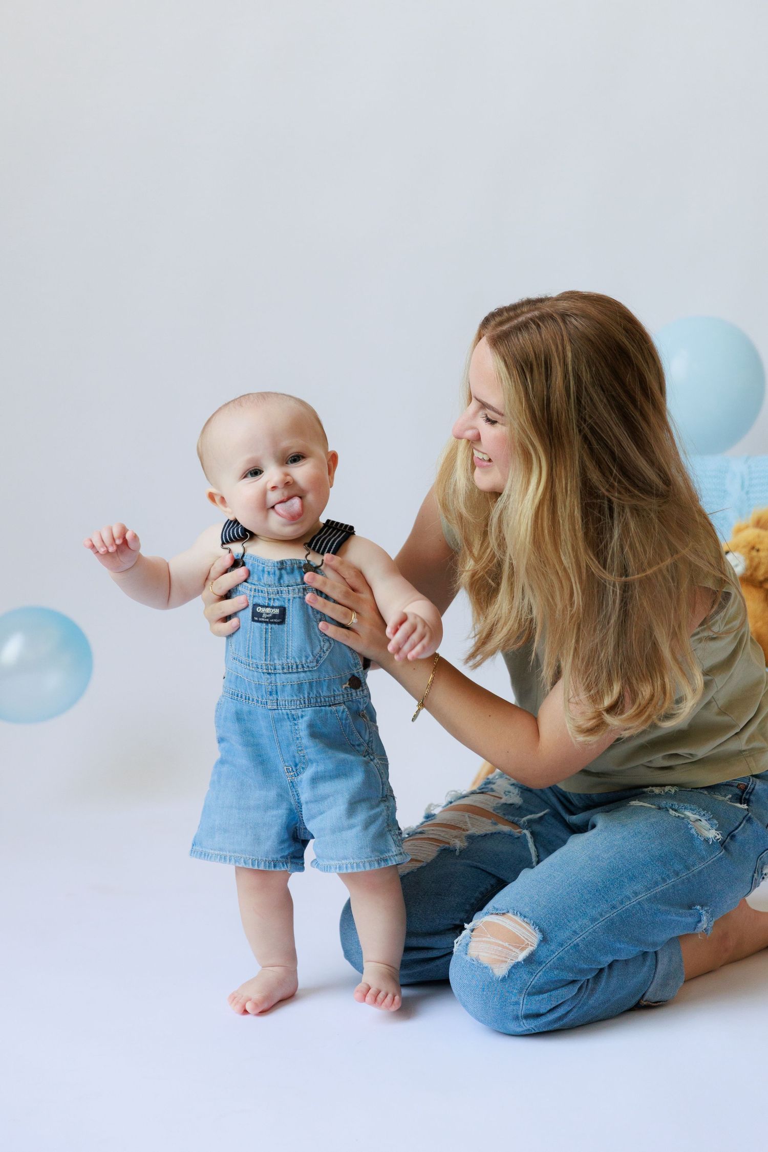 A baby in denim overalls smiles while playing with someone on a white backdrop with blue balloons.