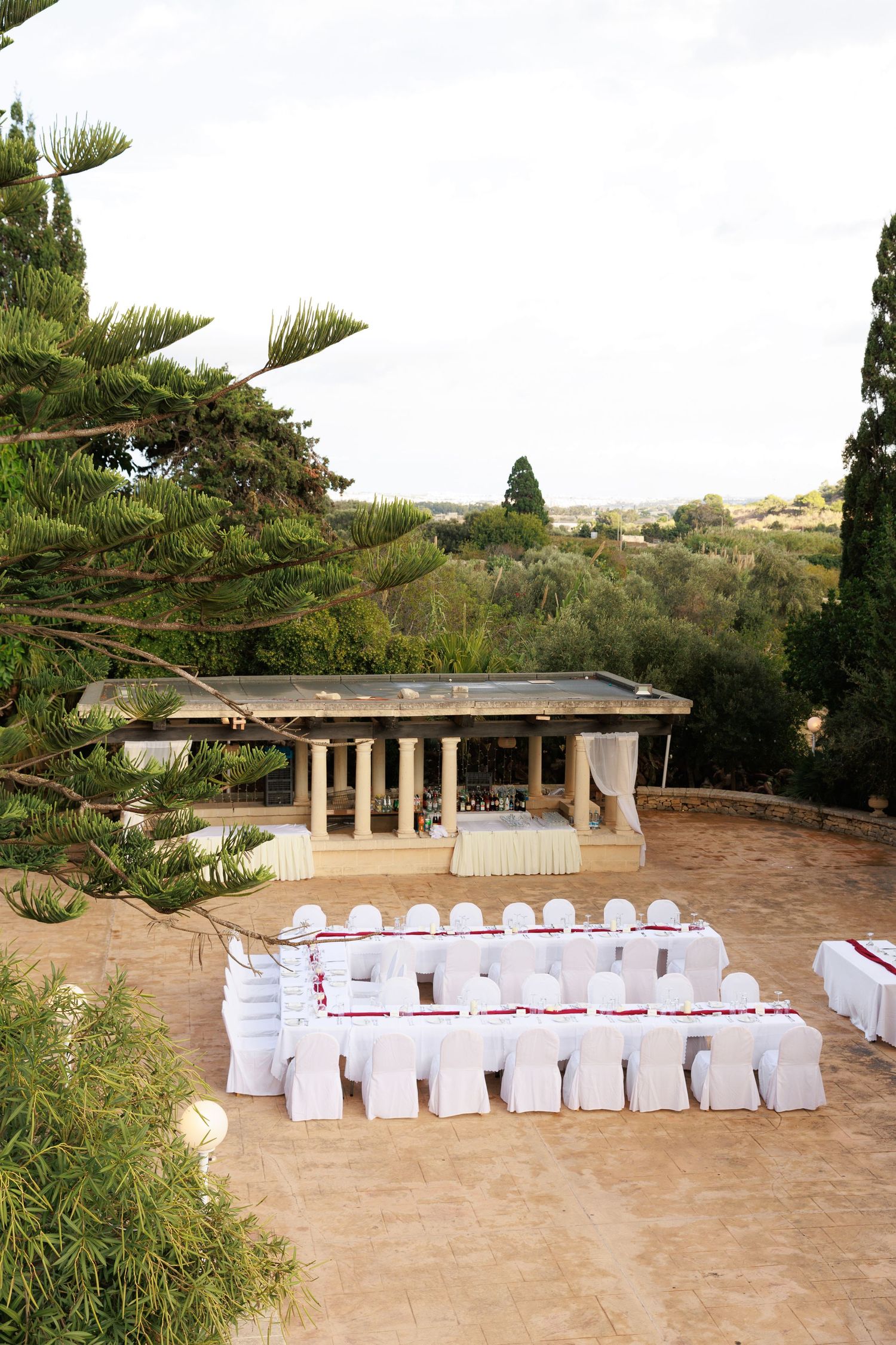 Outdoor wedding ceremony setup with white chairs and pillars arranged on a sandy terrace overlooking a scenic landscape.