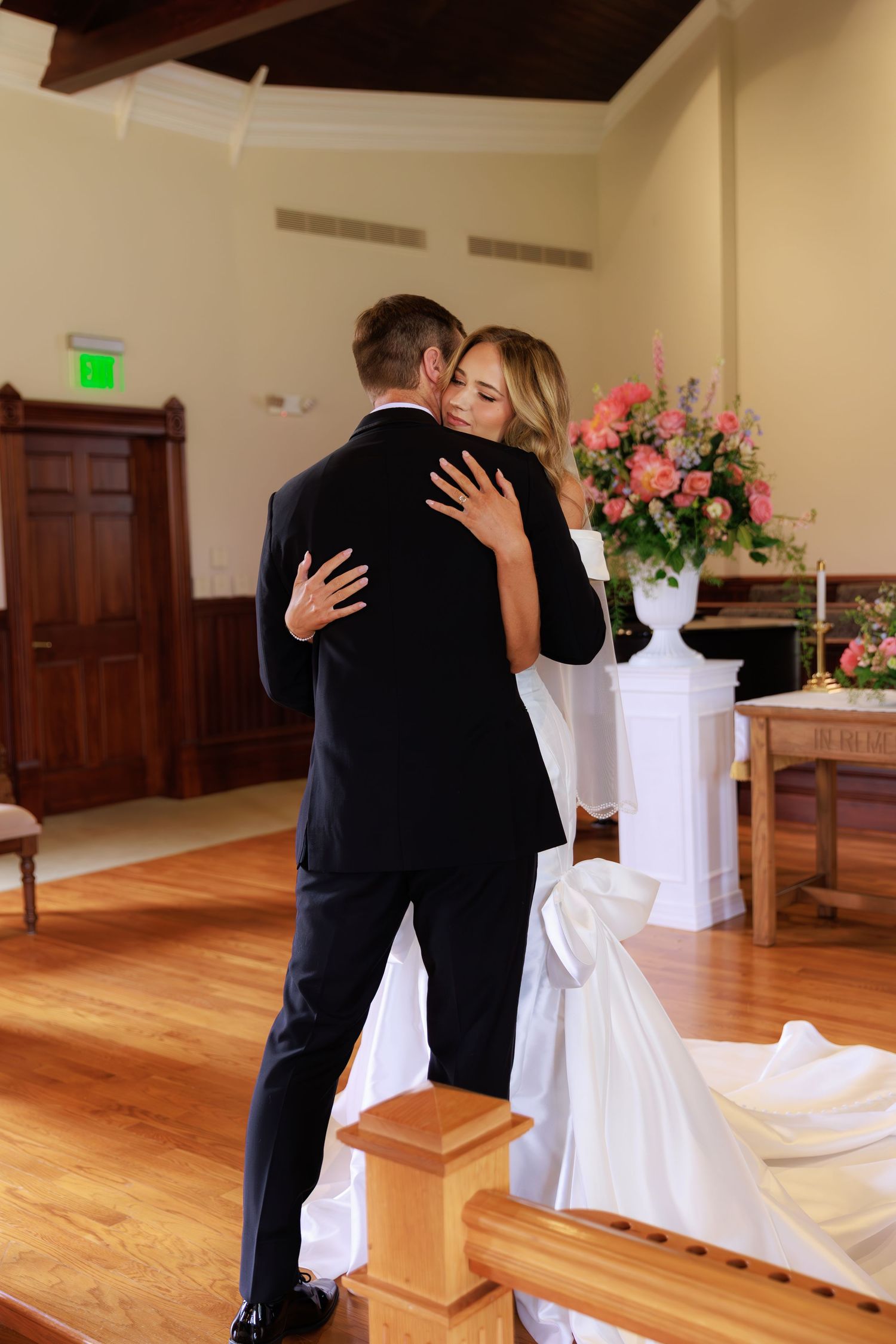 A couple embraces during their wedding ceremony in a church with hardwood floors and pink floral arrangements.