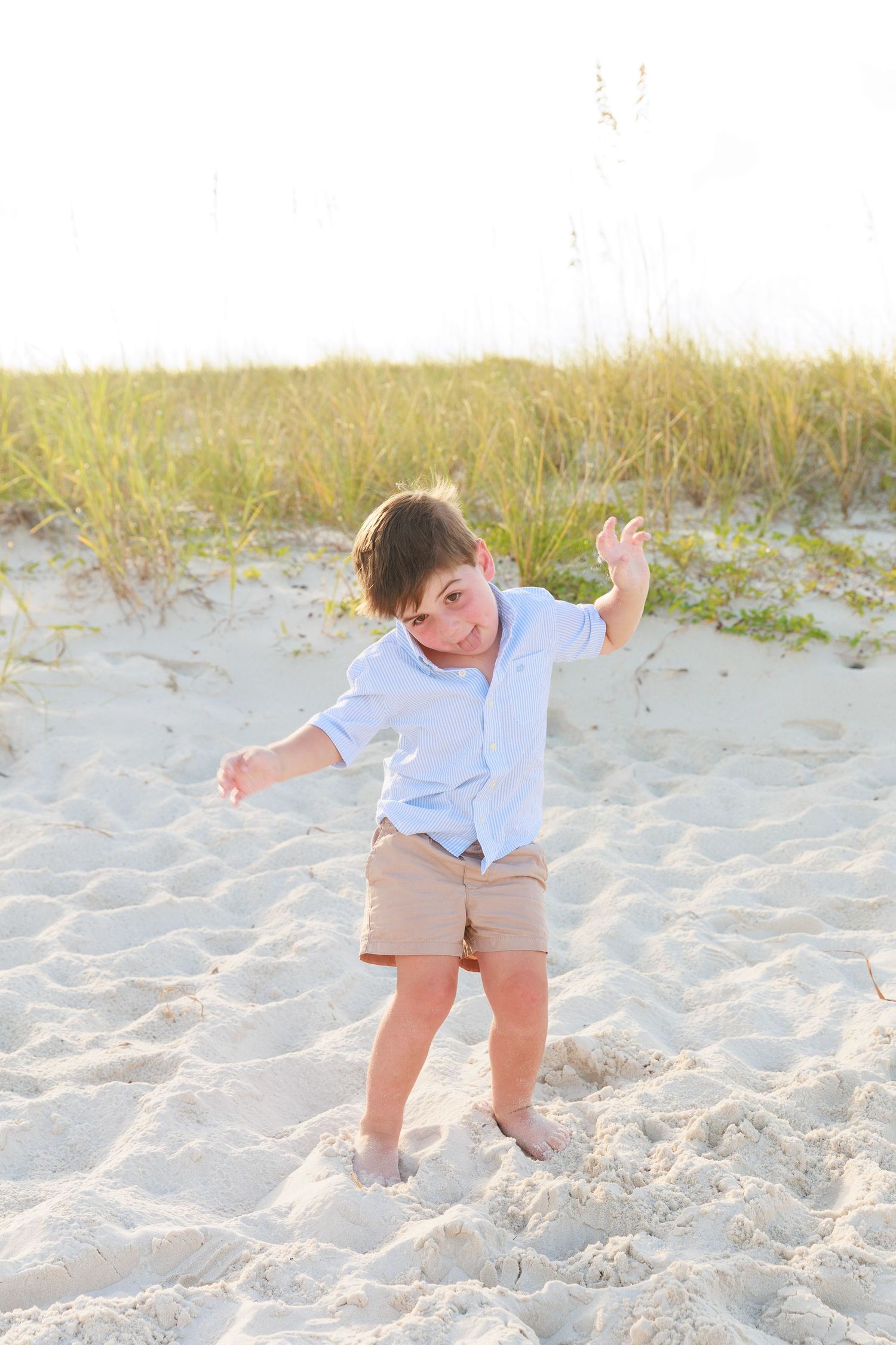 Young boy playing in beach sand dunes, wearing light blue shirt and khaki shorts on a sunny day.