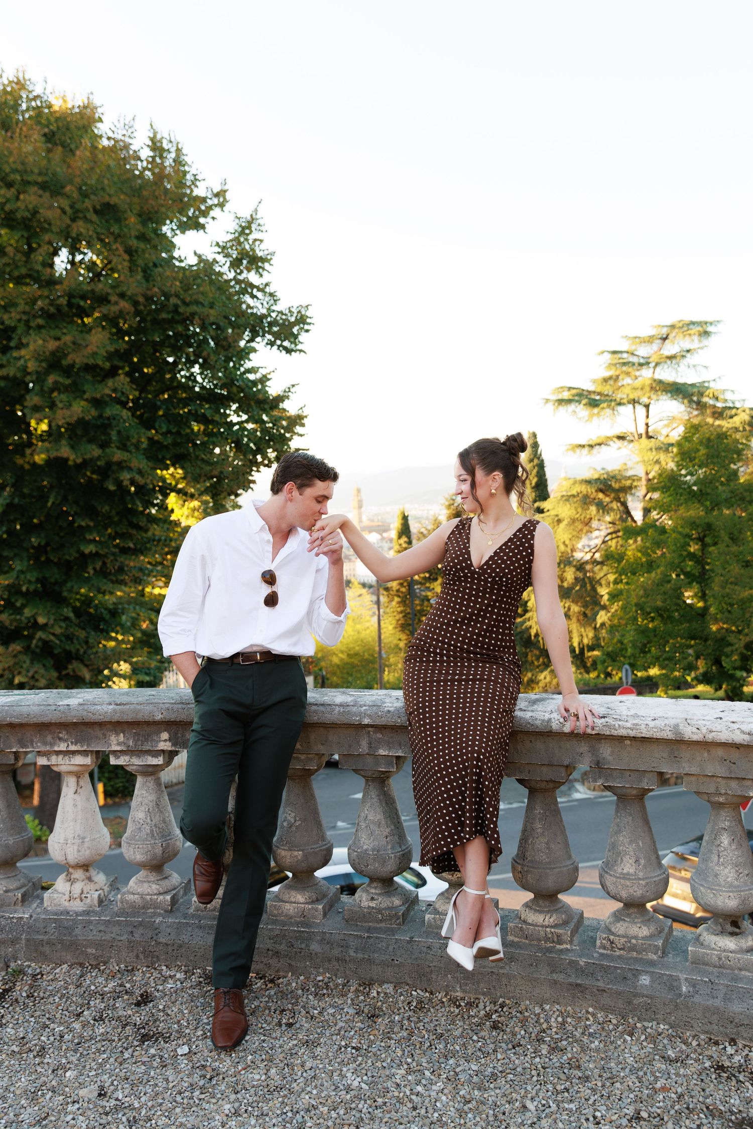 Elegant couple holding hands by ornate stone balustrade in Italian garden at sunset.
