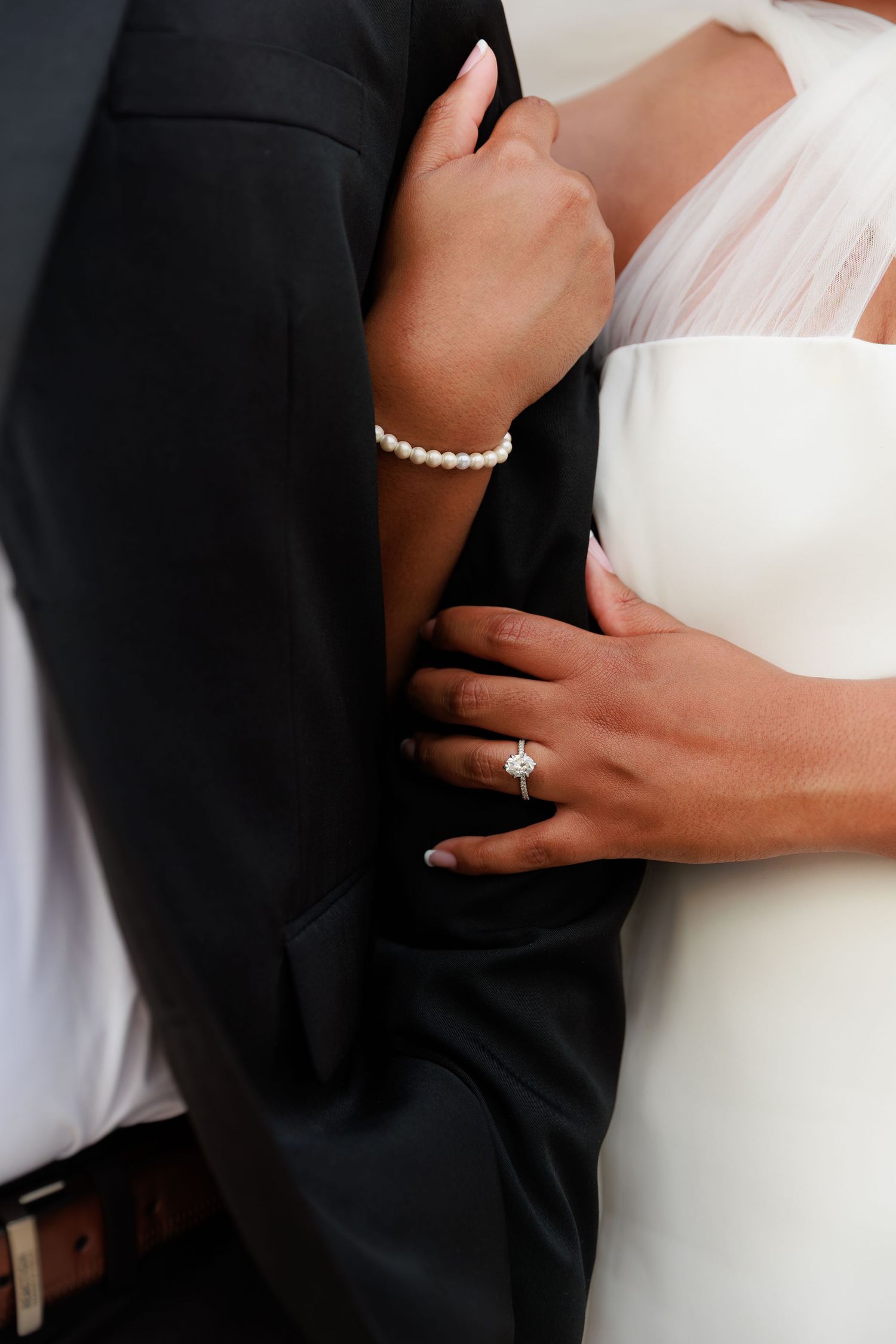 Bride wearing diamond engagement ring and pearl bracelet embracing groom in black suit at wedding ceremony.