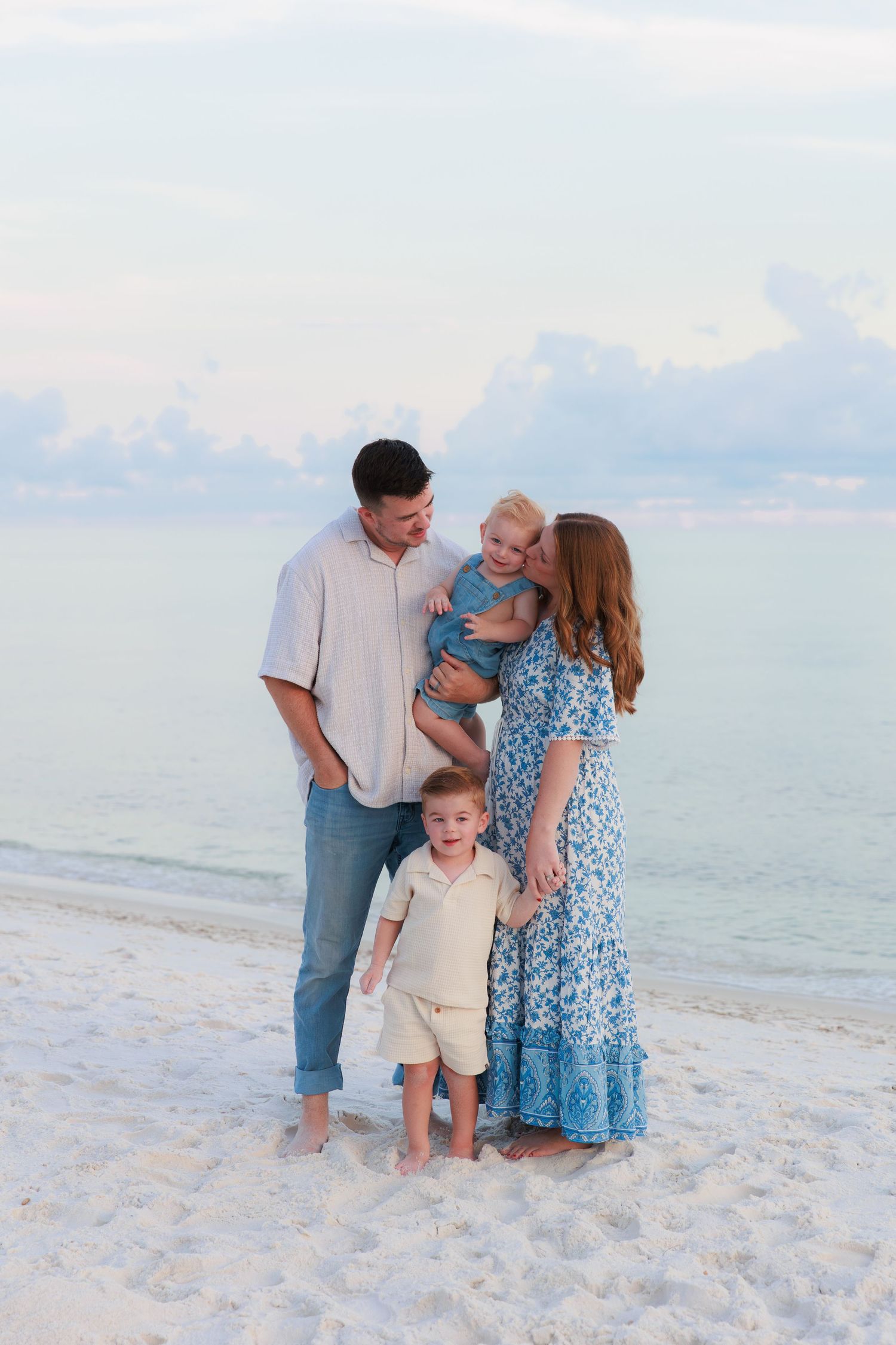 A joyful family of four stands together on a white sand beach at dusk with calm ocean waters behind them.