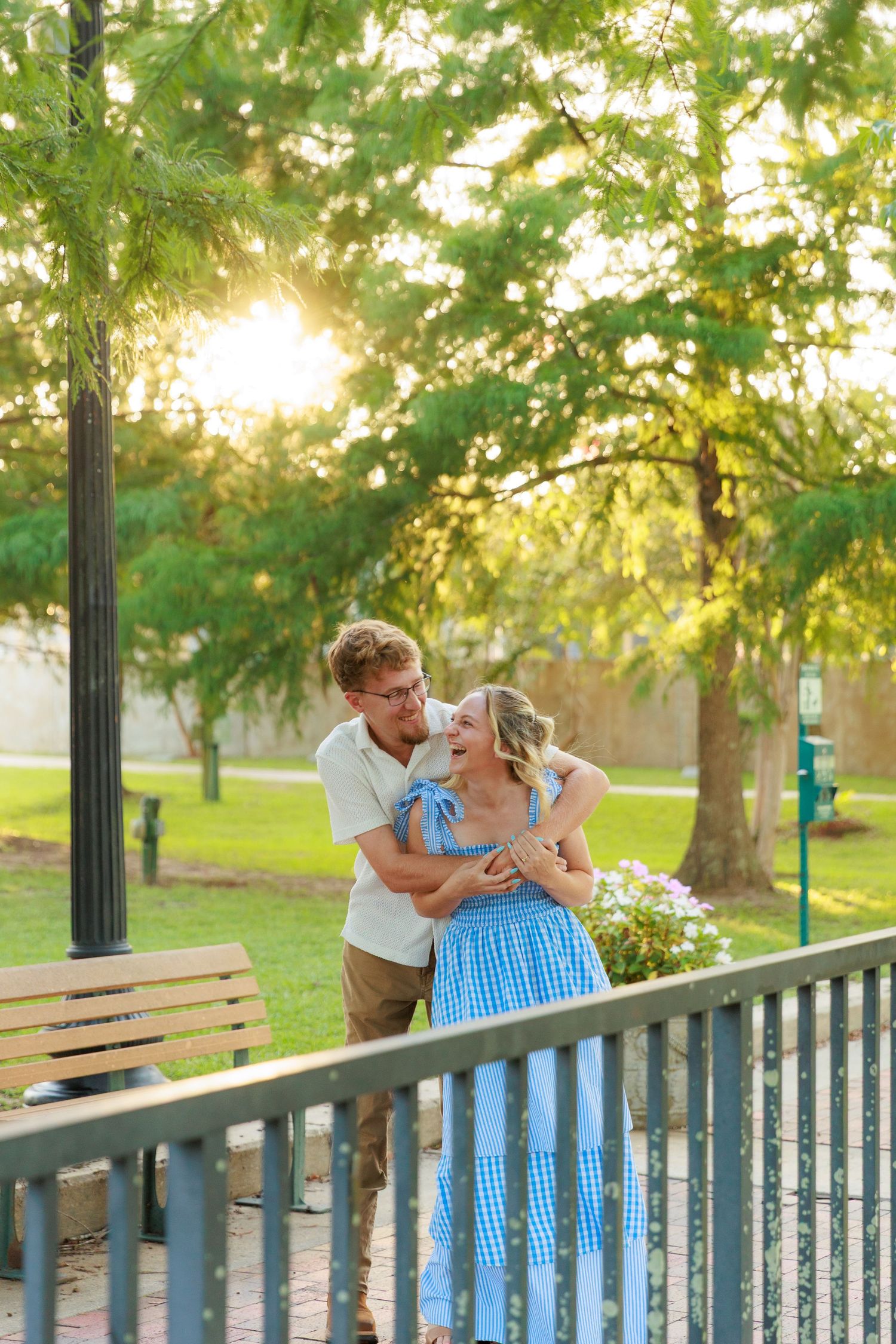 Couple embracing and smiling at sunset in a lush green park near a wooden railing and bench.