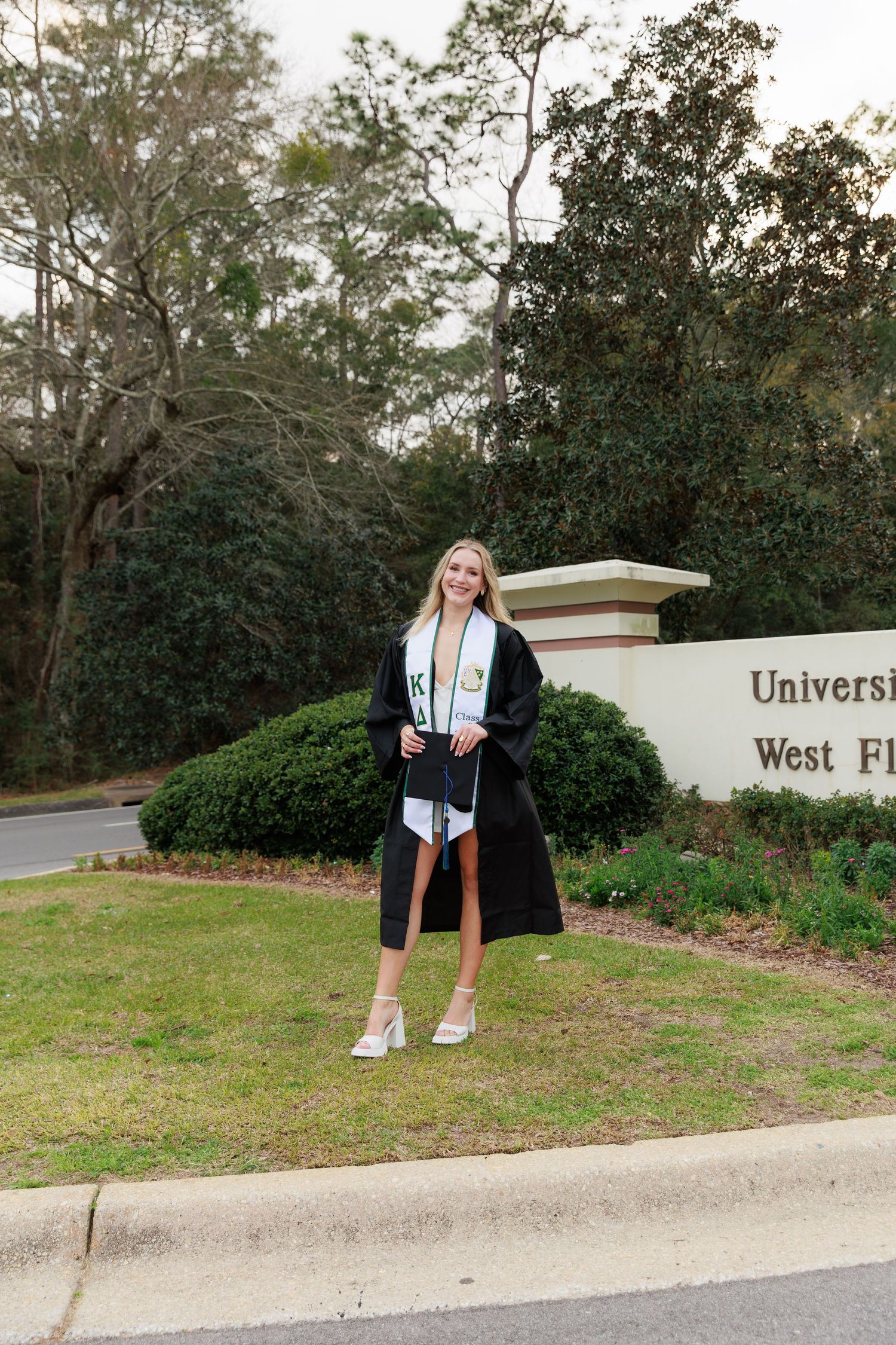 Female graduate in black robe and stole poses proudly in front of University of West Florida entrance sign.