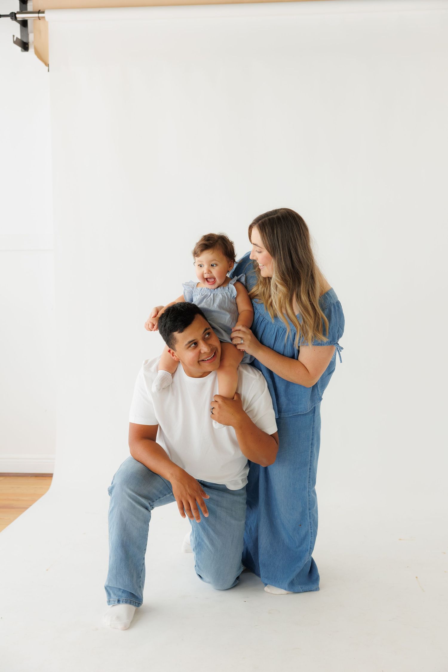 Happy family with toddler on fathers shoulders, mother in blue jumpsuit, posing in white studio.
