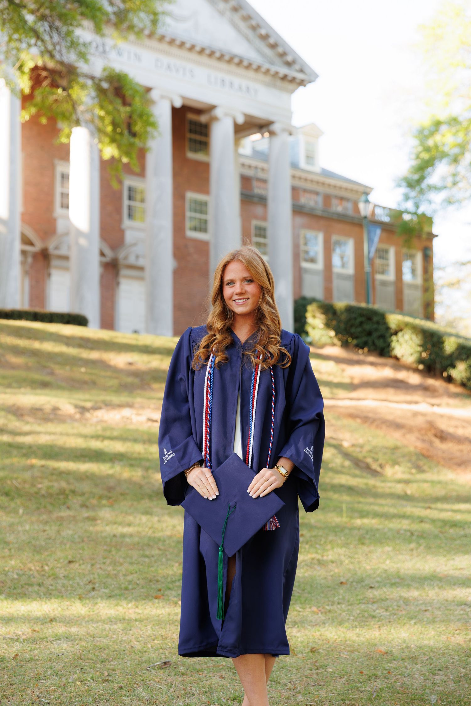 Young woman in navy blue graduation gown with honor cords stands proudly in front of a brick university library building.