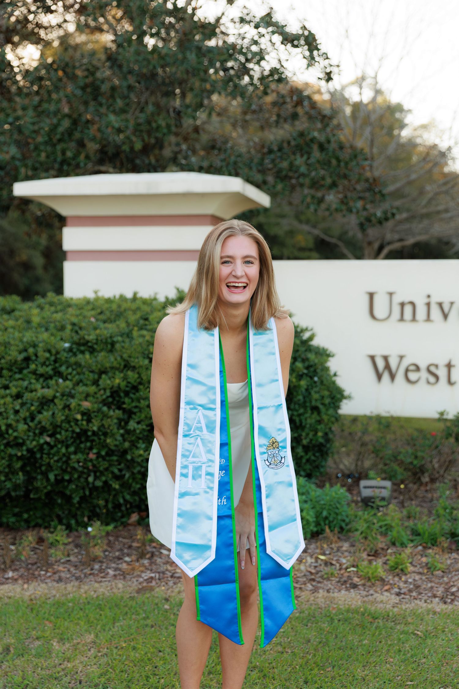 A graduate wearing a blue dress and decorated graduation stole poses for photos outdoors near university signage.