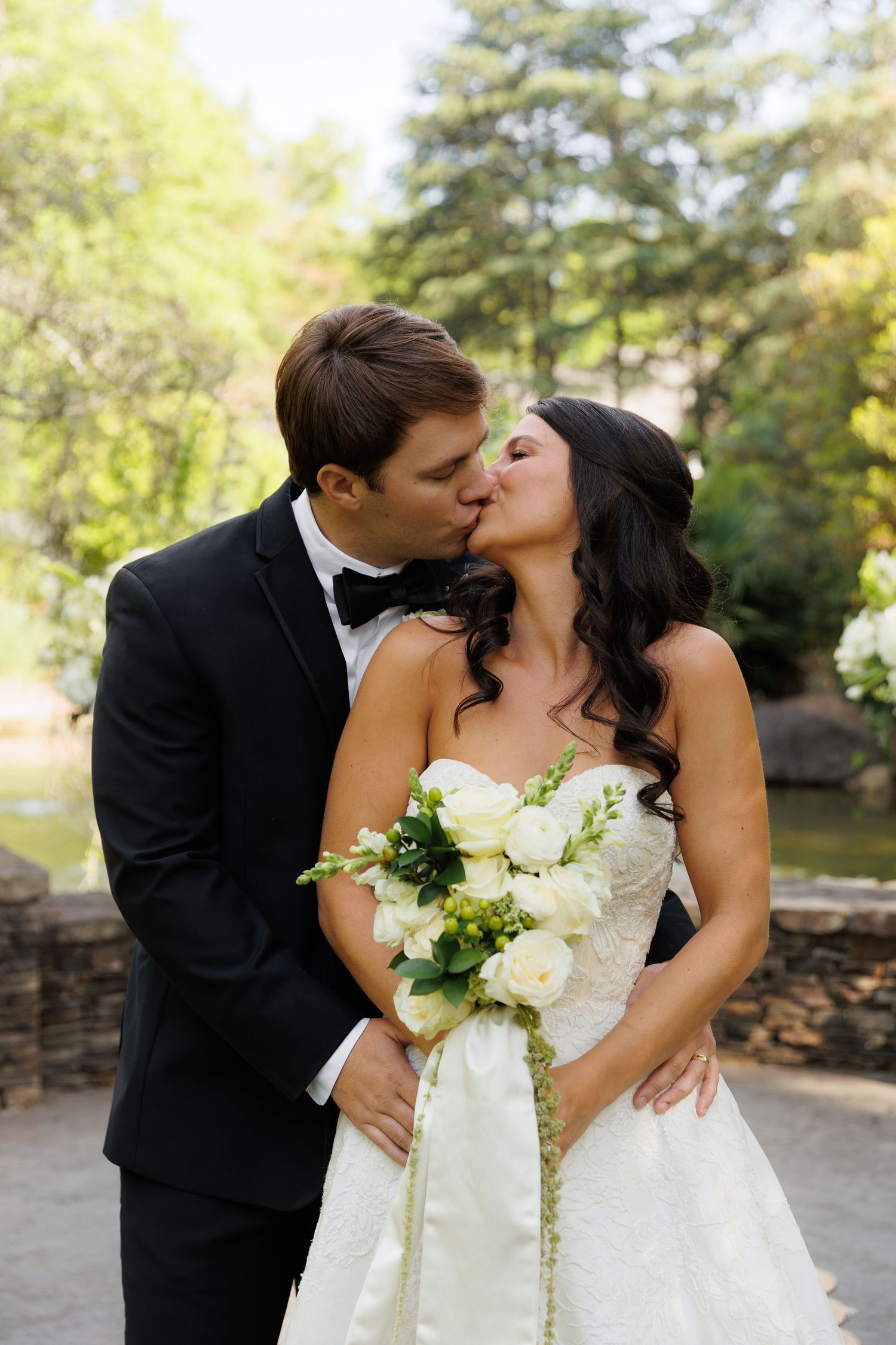 Bride and groom sharing a kiss outdoors, bride in strapless white ballgown holding white rose bouquet.