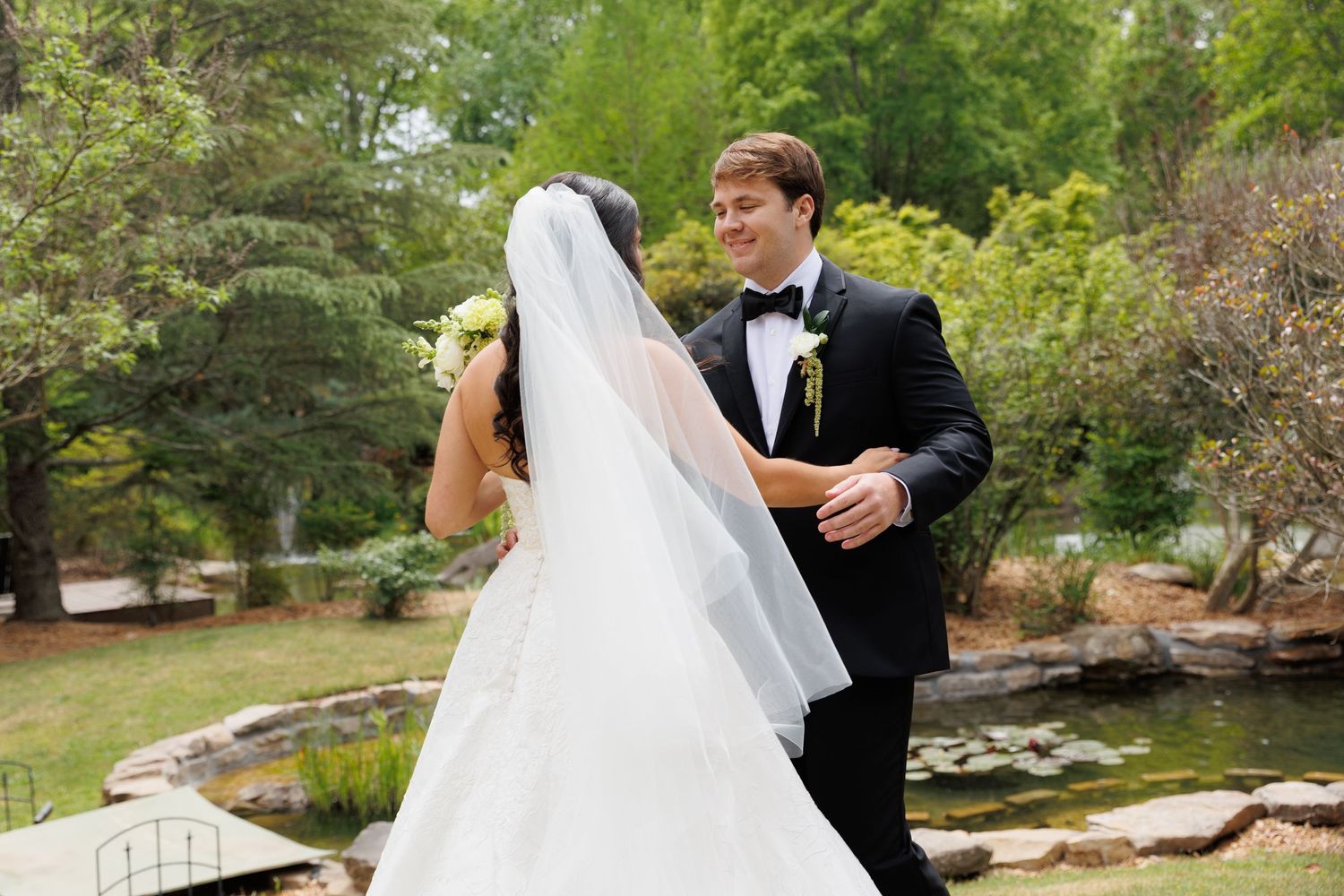 Bride in white gown and veil shares a joyful first look moment with groom in black tuxedo outdoors.