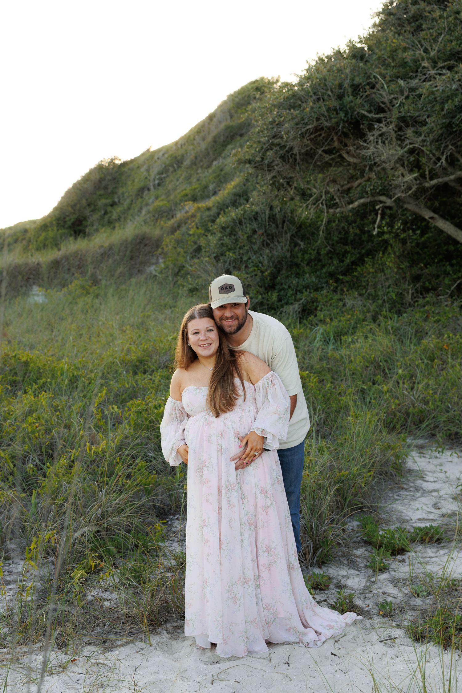 Couple poses together on sandy beach dunes, woman in floral maternity dress held by man in cap and jeans.