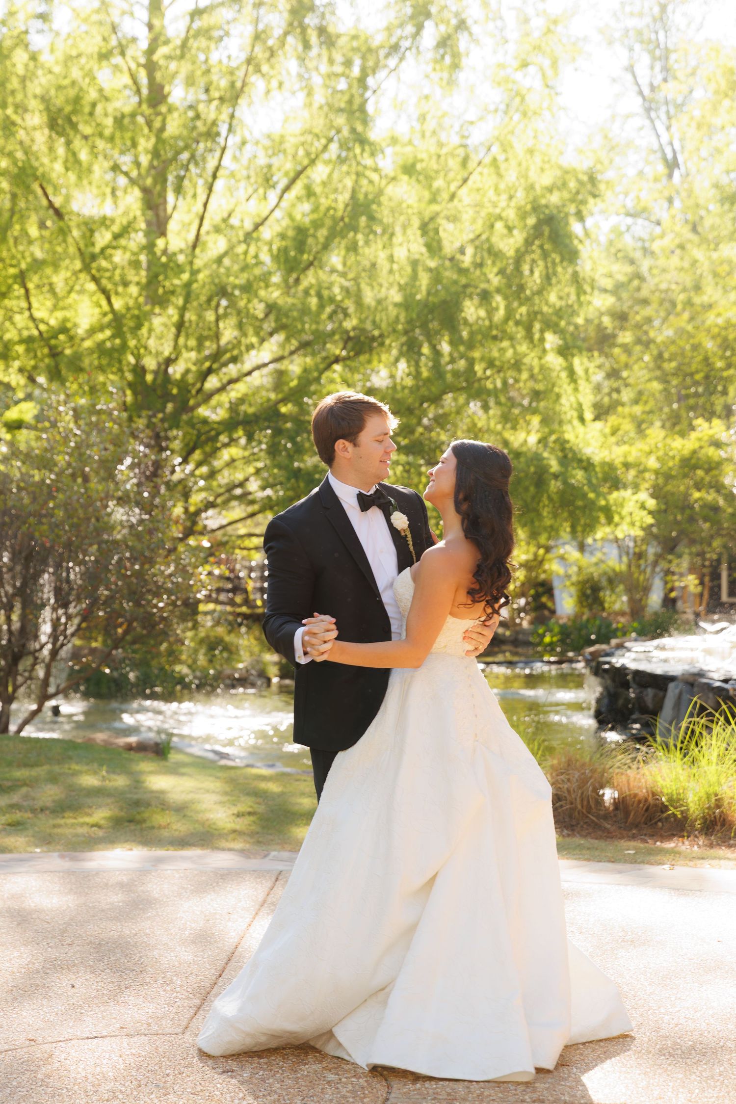 Bride and groom share a romantic dance outdoors in lush garden setting, she in white gown, he in classic black tuxedo.