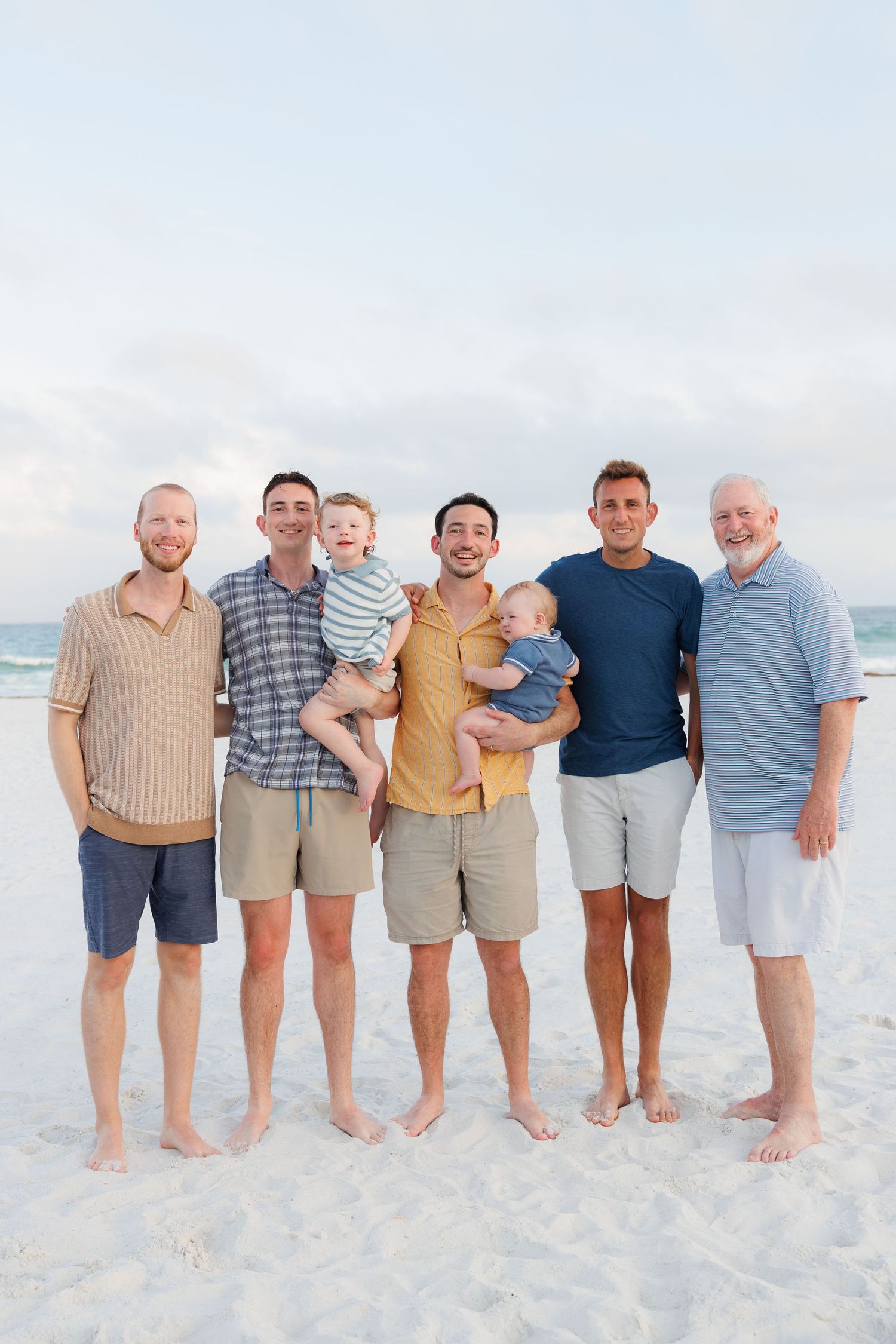 Group of men and two babies posing on a sandy beach with ocean backdrop for a family portrait.