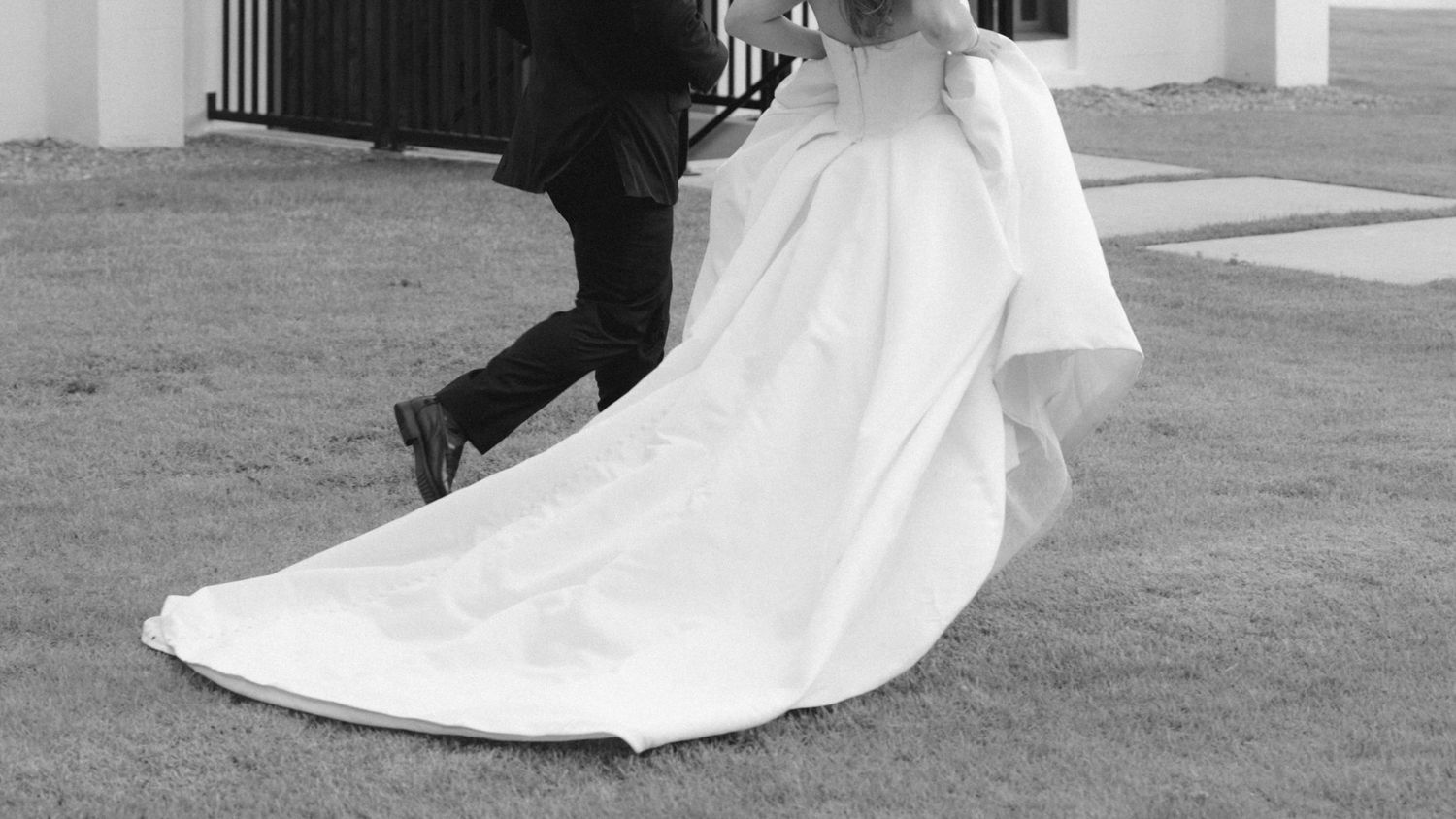Black and white photo of a bride in a flowing white wedding gown walking with a groom on a green lawn.