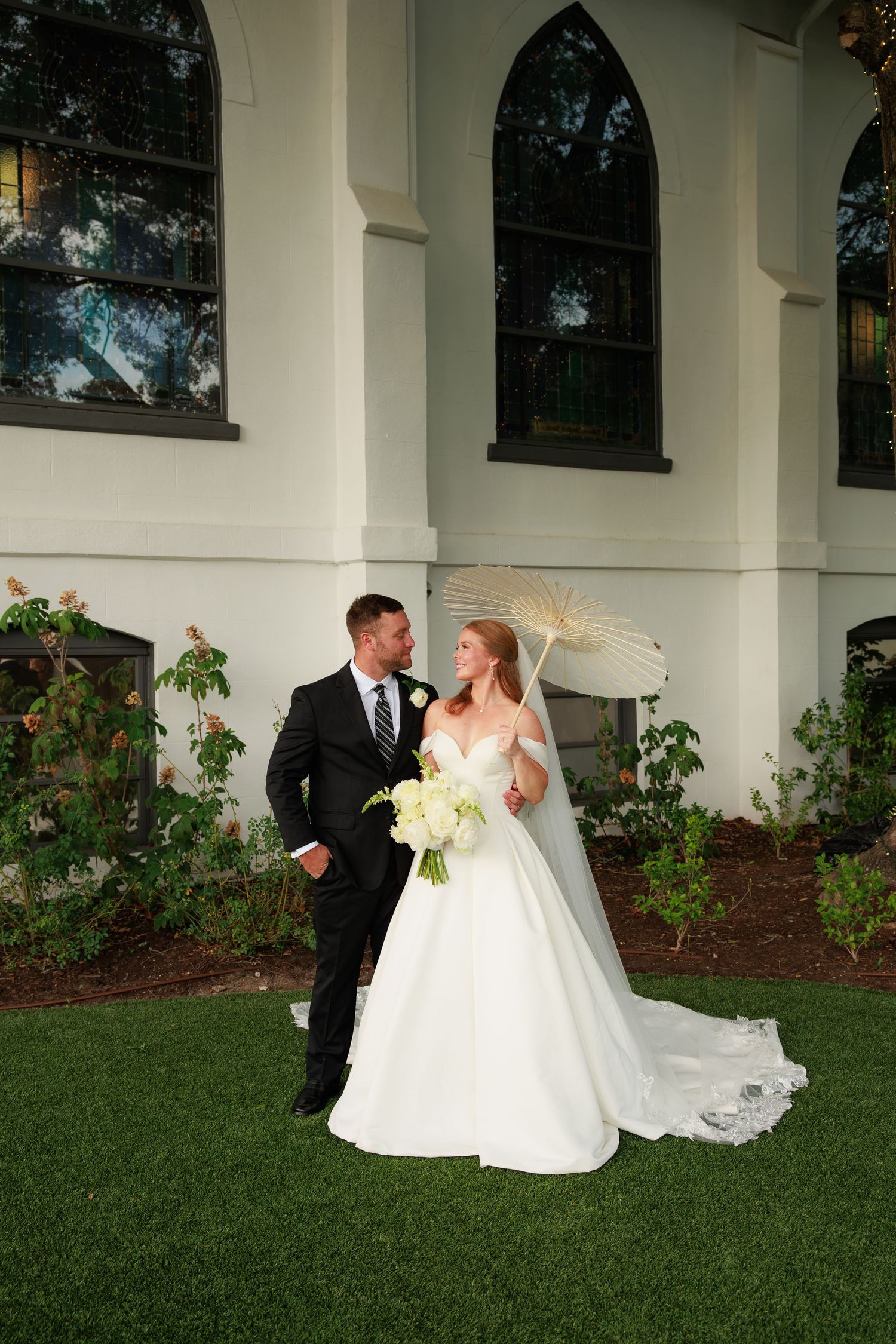 Bride in white ballgown with parasol and bouquet stands with groom in black suit outside elegant white church.