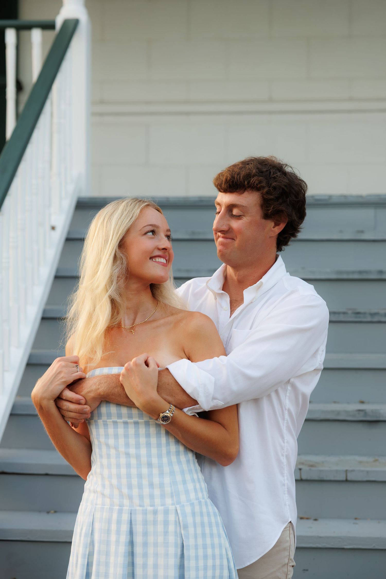 A smiling couple embraces on outdoor steps, woman in blue gingham dress, man in white shirt, sharing a tender moment.