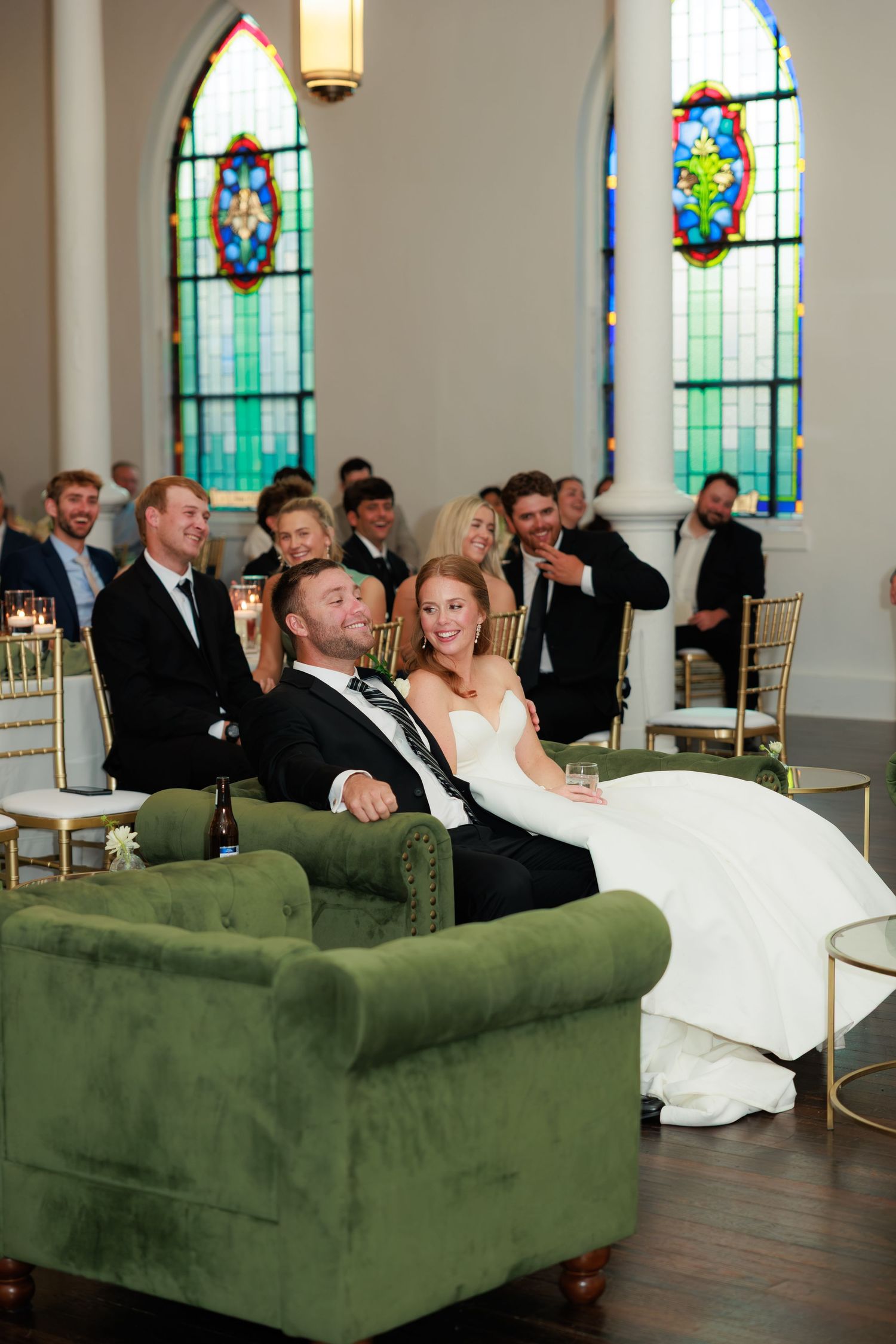 Bride and groom laughing on green velvet sofa at wedding reception in church venue with stained glass windows.